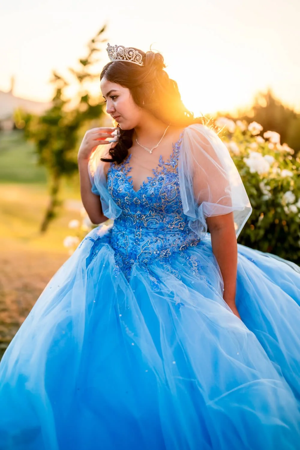 A young woman in a blue princess dress with lace and beadwork, wearing a tiara, standing outdoors during sunset, with greenery and flowers in the background.