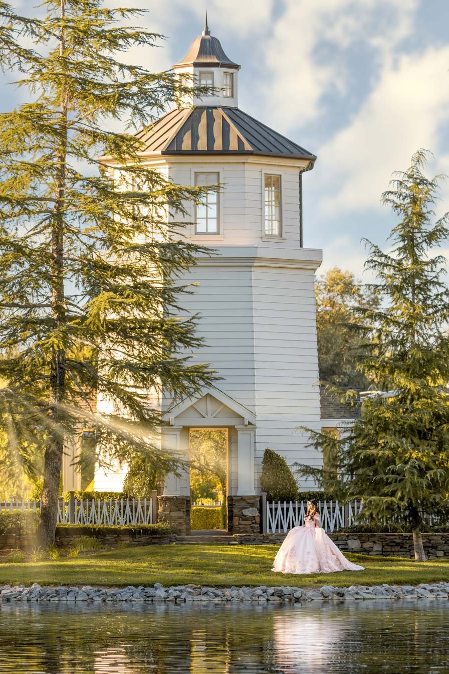 A white wooden tower with a steep roof and windows, surrounded by pine trees, sunlight, and a girl in a pink dress standing near a small lake.