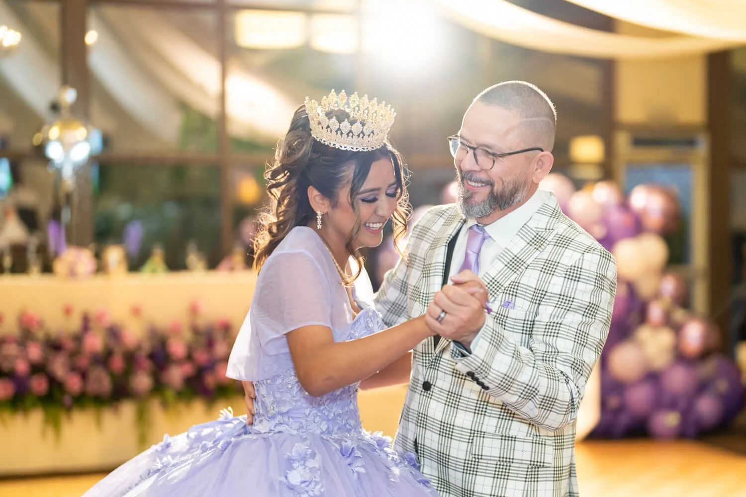 A bride wearing a crown and a white wedding gown dancing with a groom in a plaid suit at a wedding reception, with pink and purple floral decorations in the background.