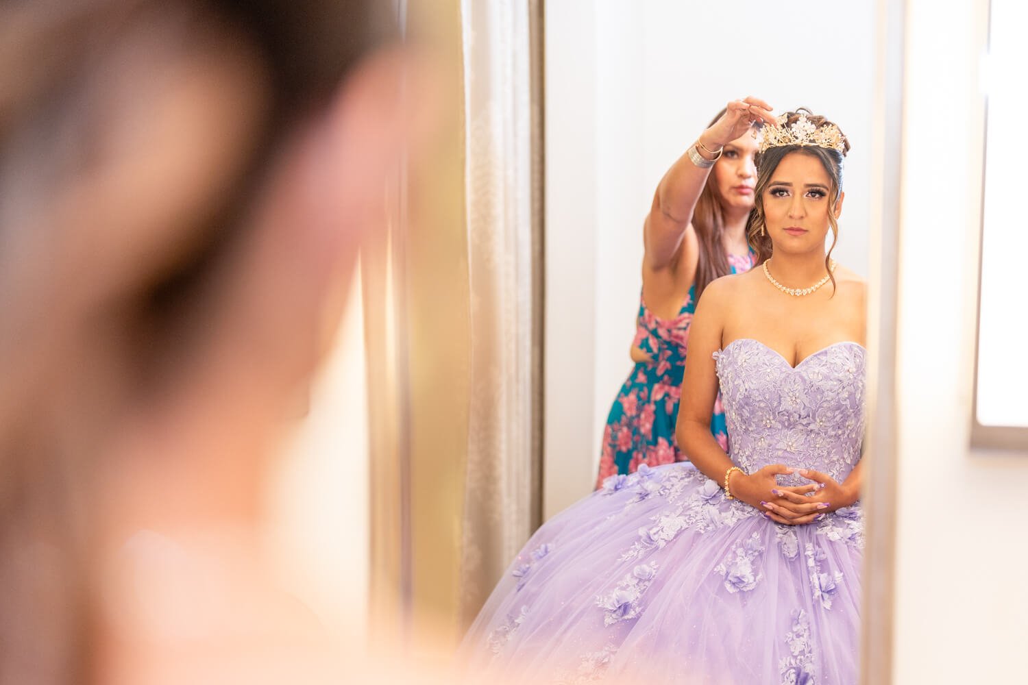 Young woman in a lavender bridal gown looking in mirror, with woman adjusting tiara in the background.