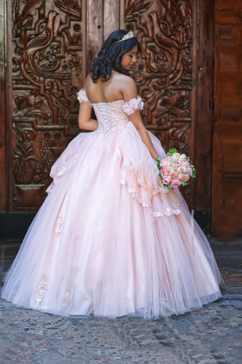 A woman in a pink ball gown with floral details and lace, wearing a tiara, holding a pink and white bouquet, standing in front of intricately carved wooden doors.