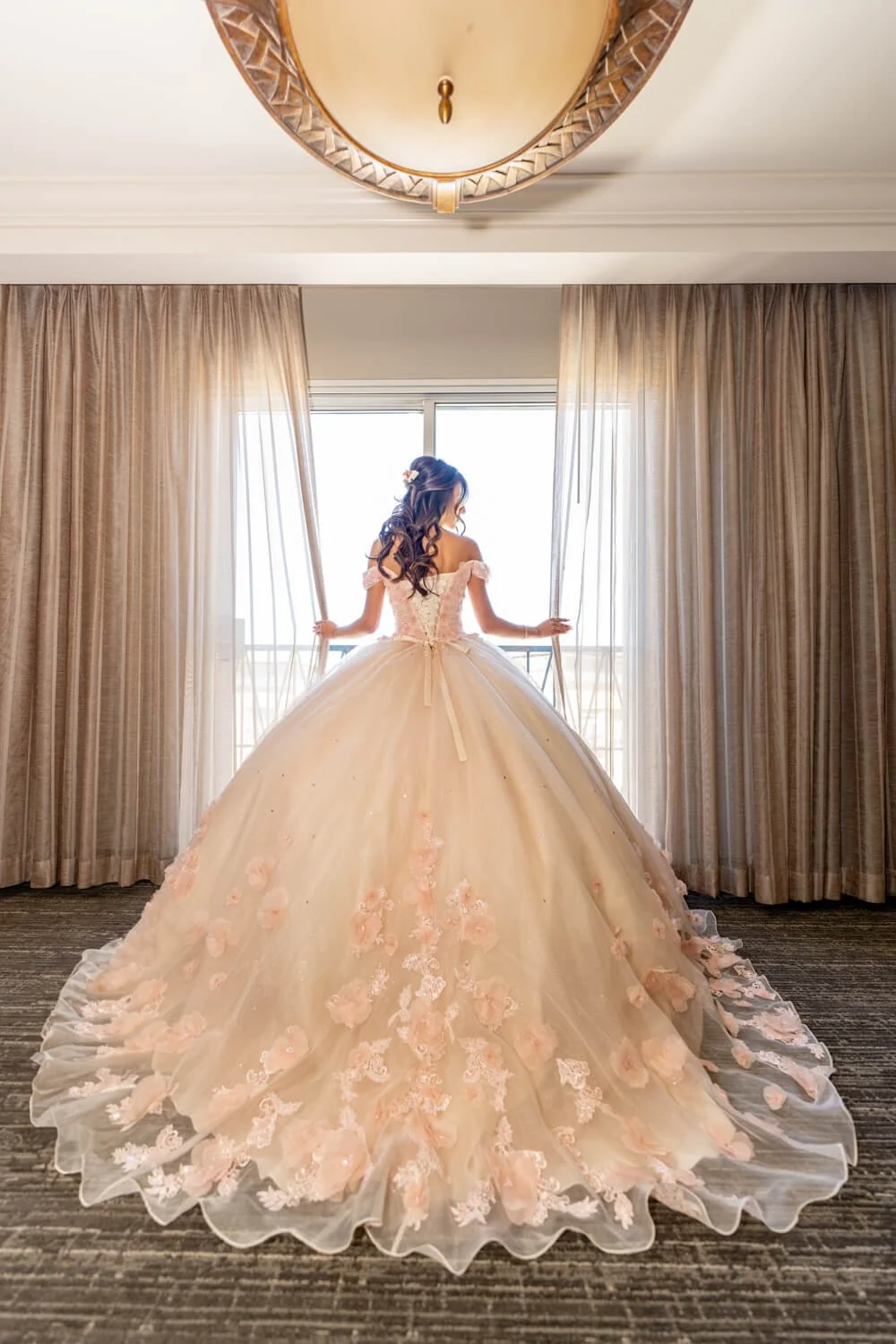 A woman in a pink wedding dress with floral embroidery stands in front of a large window with sheer curtains, looking outside, with her back to the camera.