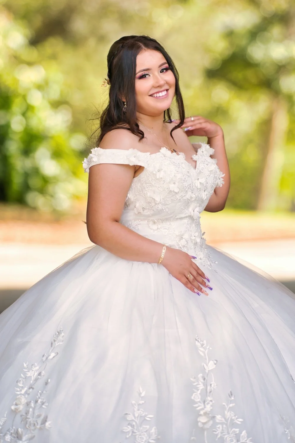 A young woman in a white wedding dress with floral appliqué and off-shoulder design, standing outdoors with a blurred green background. She has dark hair styled in loose curls, is smiling, and wearing jewelry.
