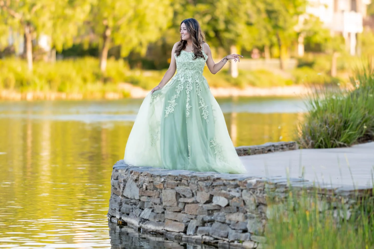 Young woman in a light green dress with floral embroidery standing on a stone edge near a body of water, surrounded by green trees.