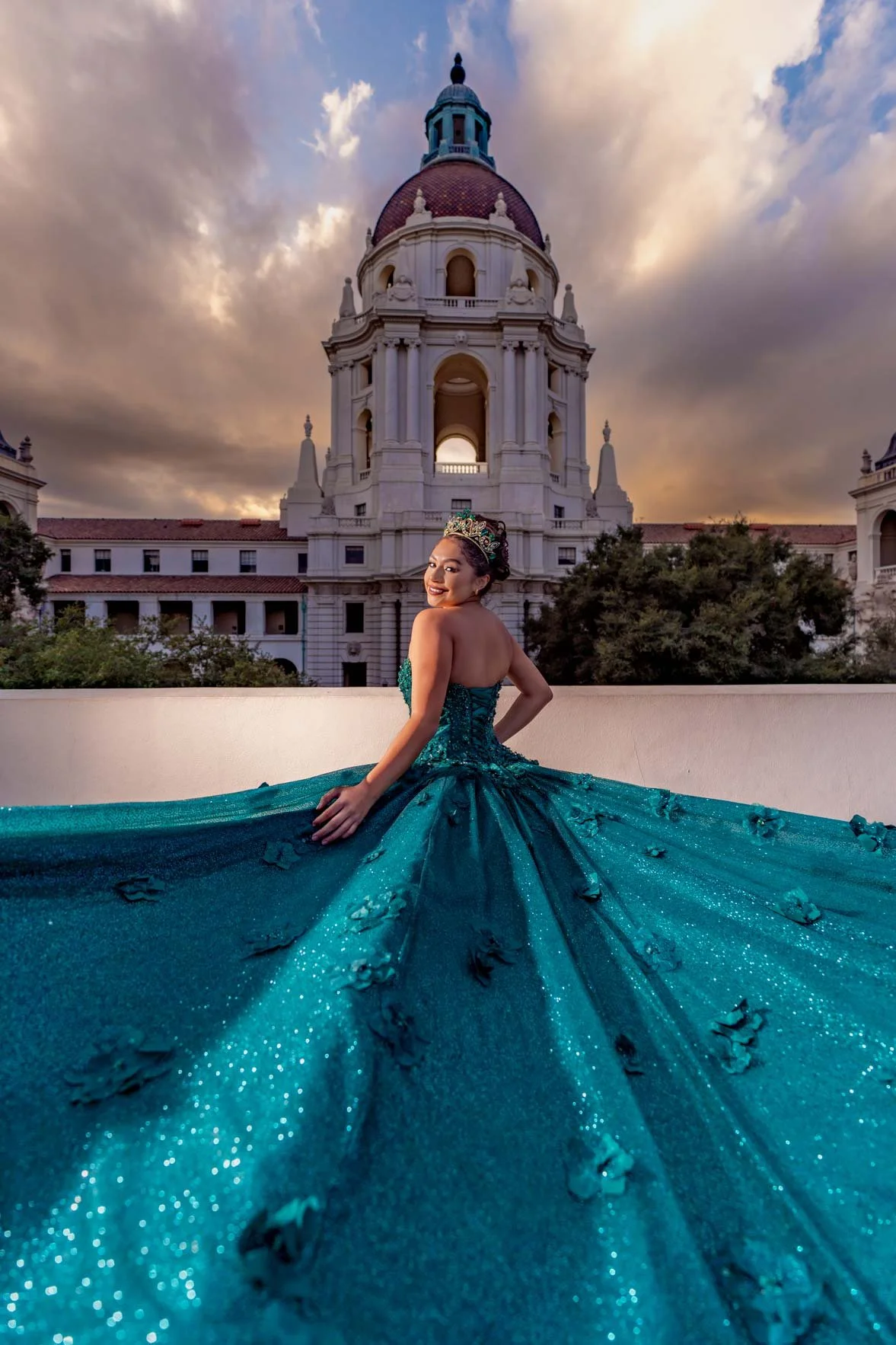 A woman in a teal, glittery gown and crown is smiling and sitting on a terrace with an ornate, historic building with a large dome and a clock tower in the background during sunset.