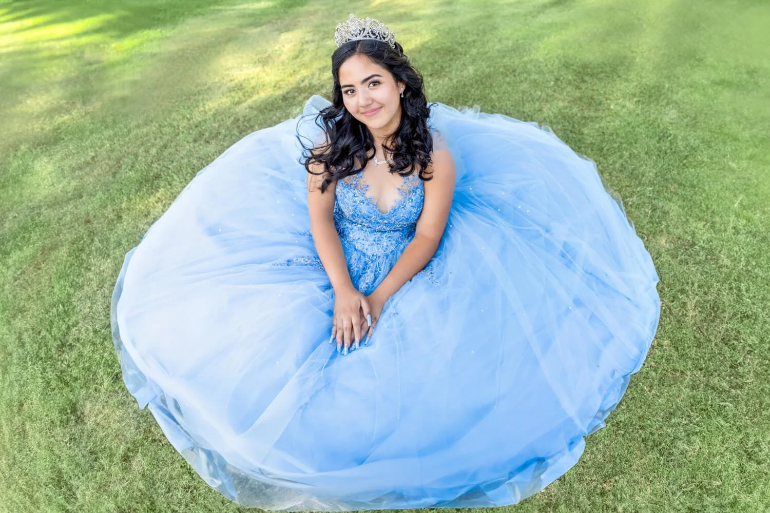 A young woman in a blue ball gown and tiara sitting on grass, smiling at the camera.
