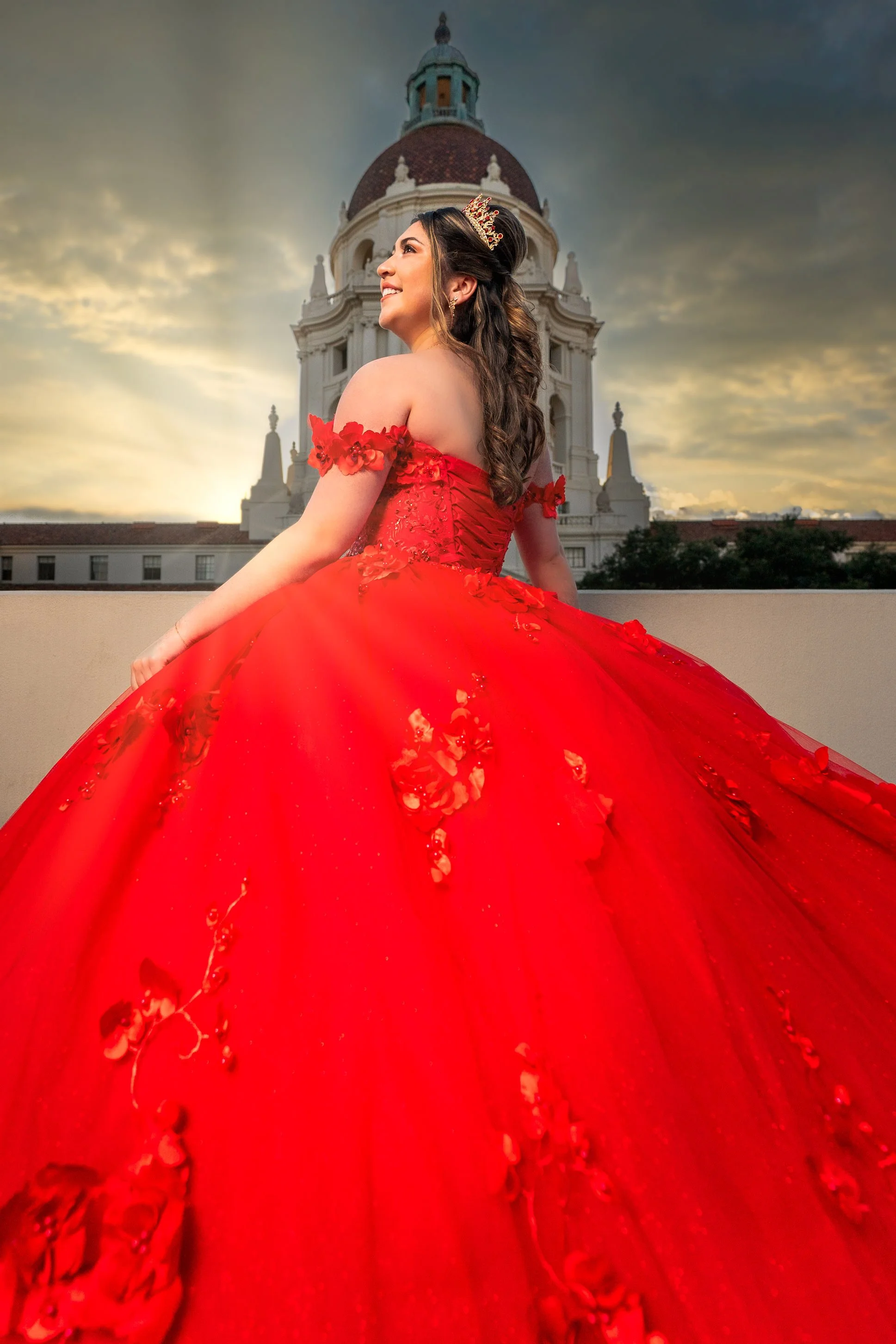 A woman in an elegant red ball gown with floral embellishments, wearing a crown, stands outdoors at sunset with a historic building with a dome and towers in the background.