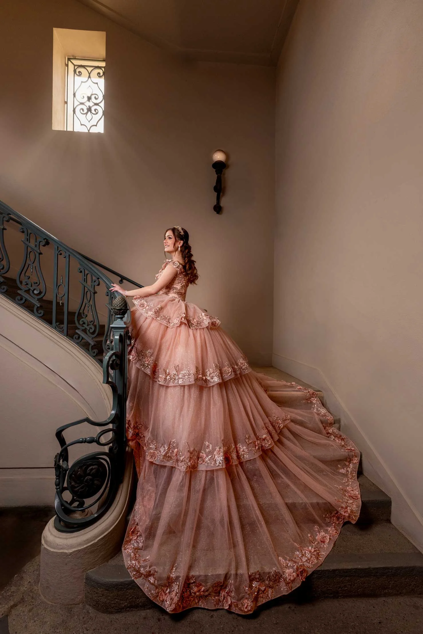 A woman wearing a pink, multi-layered, embroidered gown with a long train standing on a staircase, holding the railing, and smiling inside a building with beige walls and a small window.