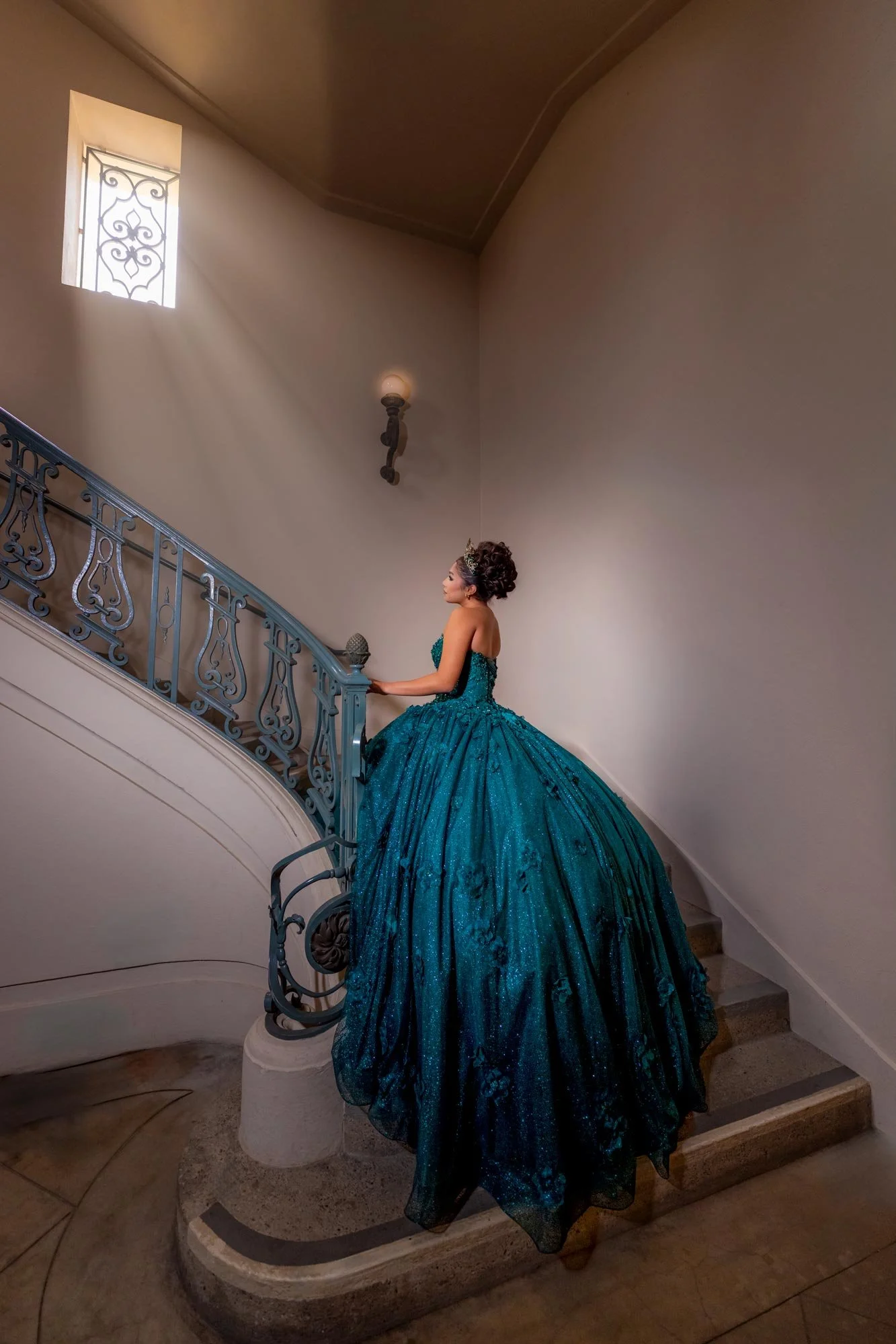 A woman in a teal, strapless, ball gown with a full skirt, ascending a curved staircase inside a building with beige walls and a small window with decorative ironwork.