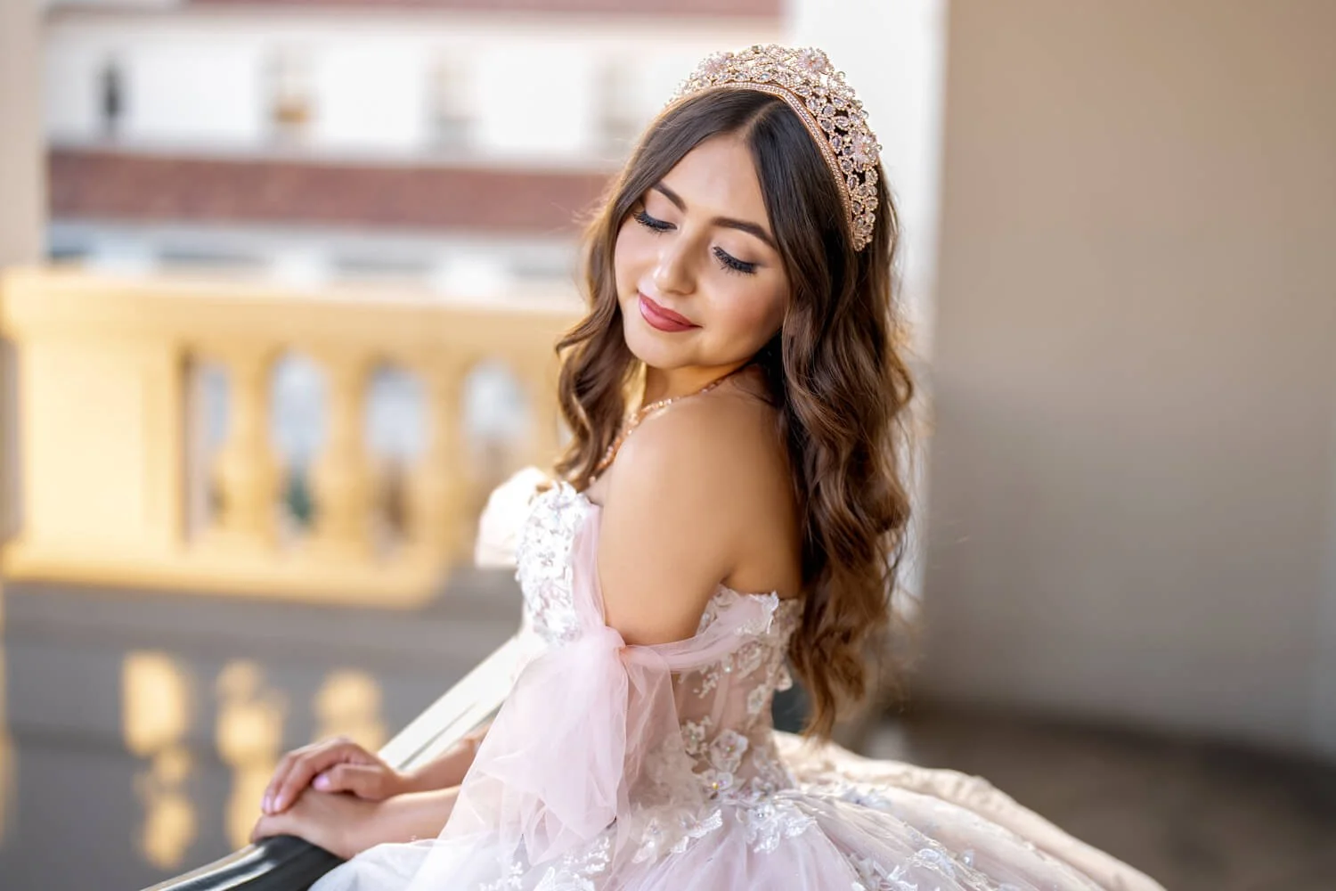 A young woman with long brown curly hair wearing a pink off-shoulder wedding dress with lace details, a pearl headband, and a pearl necklace, sitting outdoors with a blurred background of a building and yellow railing.