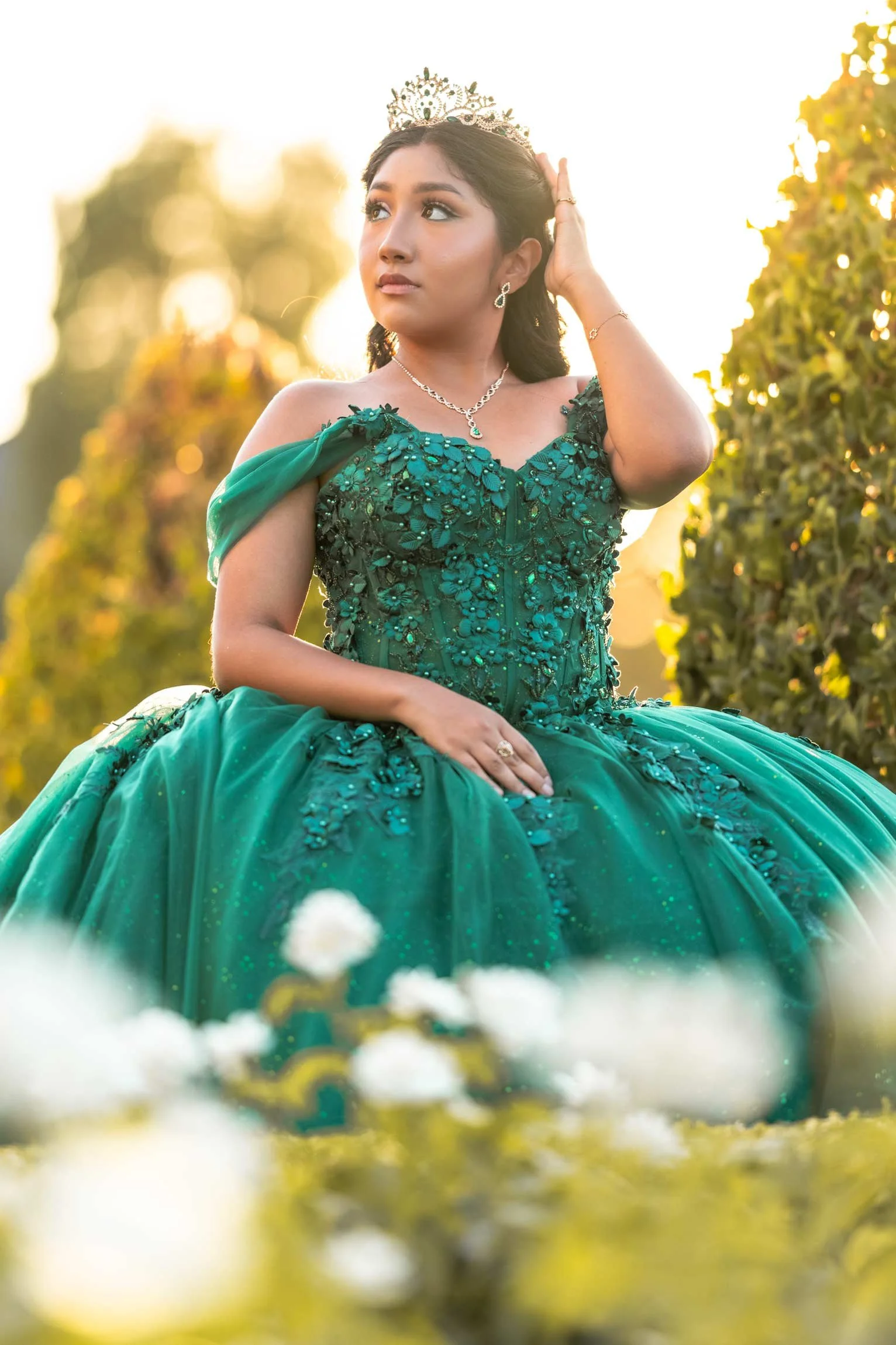A woman in a green gown and tiara sitting outdoors, with sunlight in the background, touching her hair and wearing jewelry.
