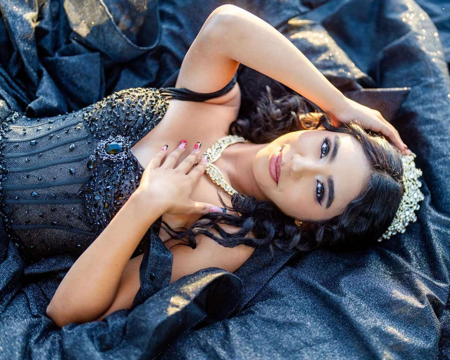 A woman with long black hair and blue eyes lying on a dark, shiny fabric. She is wearing a black, beaded dress with a large black gemstone jewel on her side, and a pearl necklace. Her hand is on her chest and her other hand is resting on her head, wi