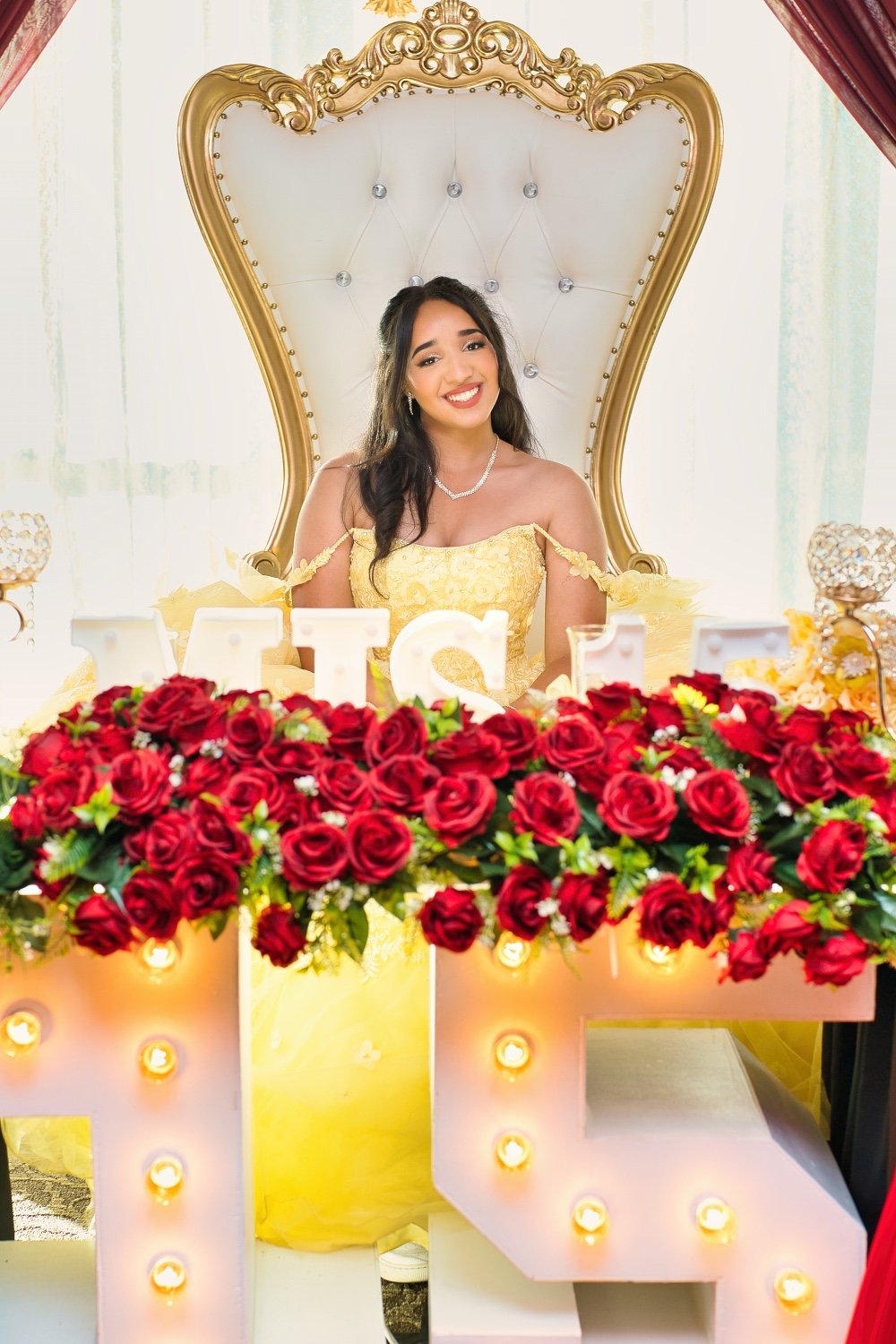 Young woman in a yellow dress seated on a large ornate white and gold throne at a celebration, with red roses and illuminated letters in front of her.