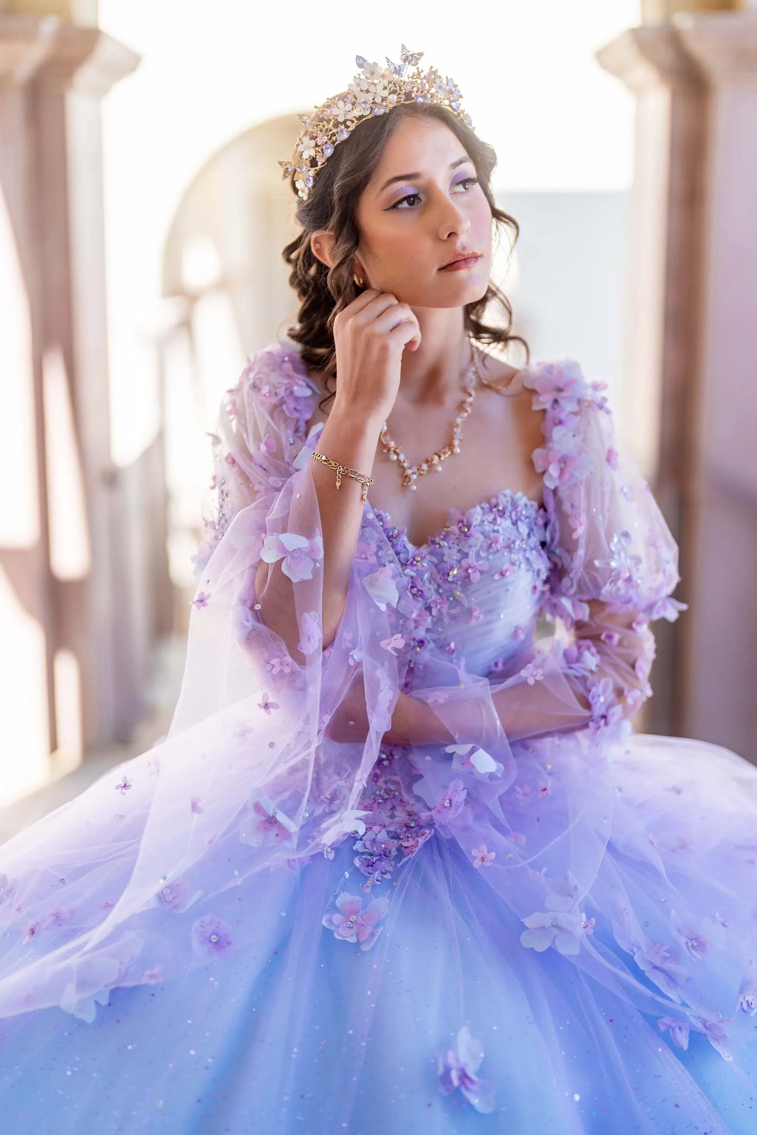 Young woman in a lavender gown with floral embellishments, wearing a tiara and jewelry, sitting in a well-lit room.