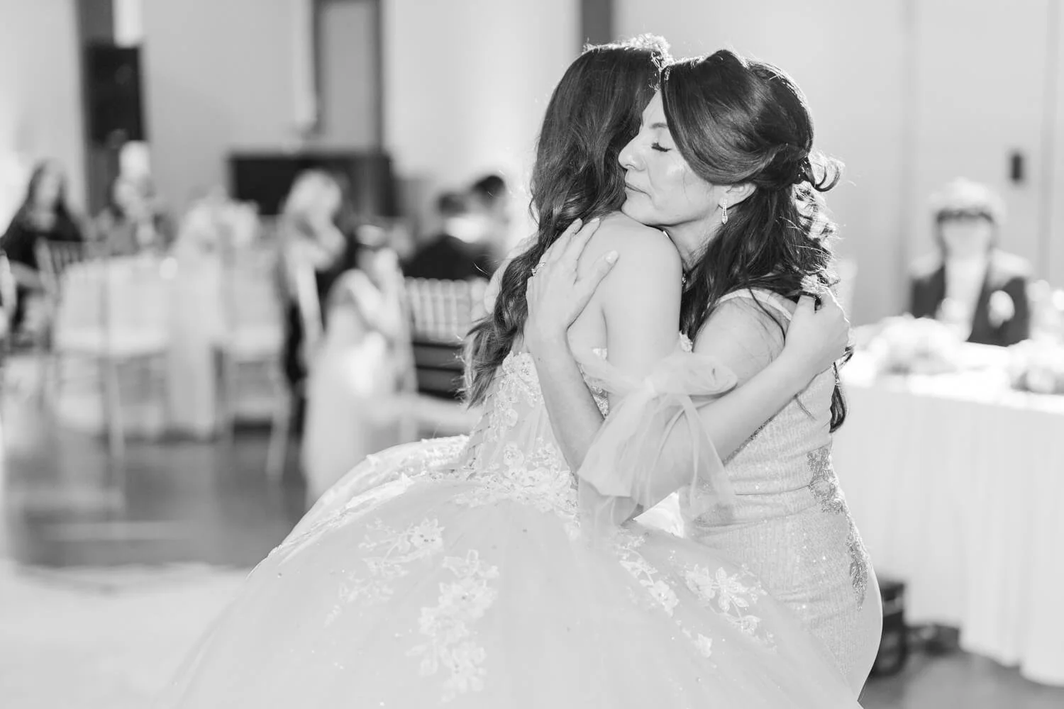 Two women embracing, one in a wedding dress, in a reception hall with guests seated at tables in the background.