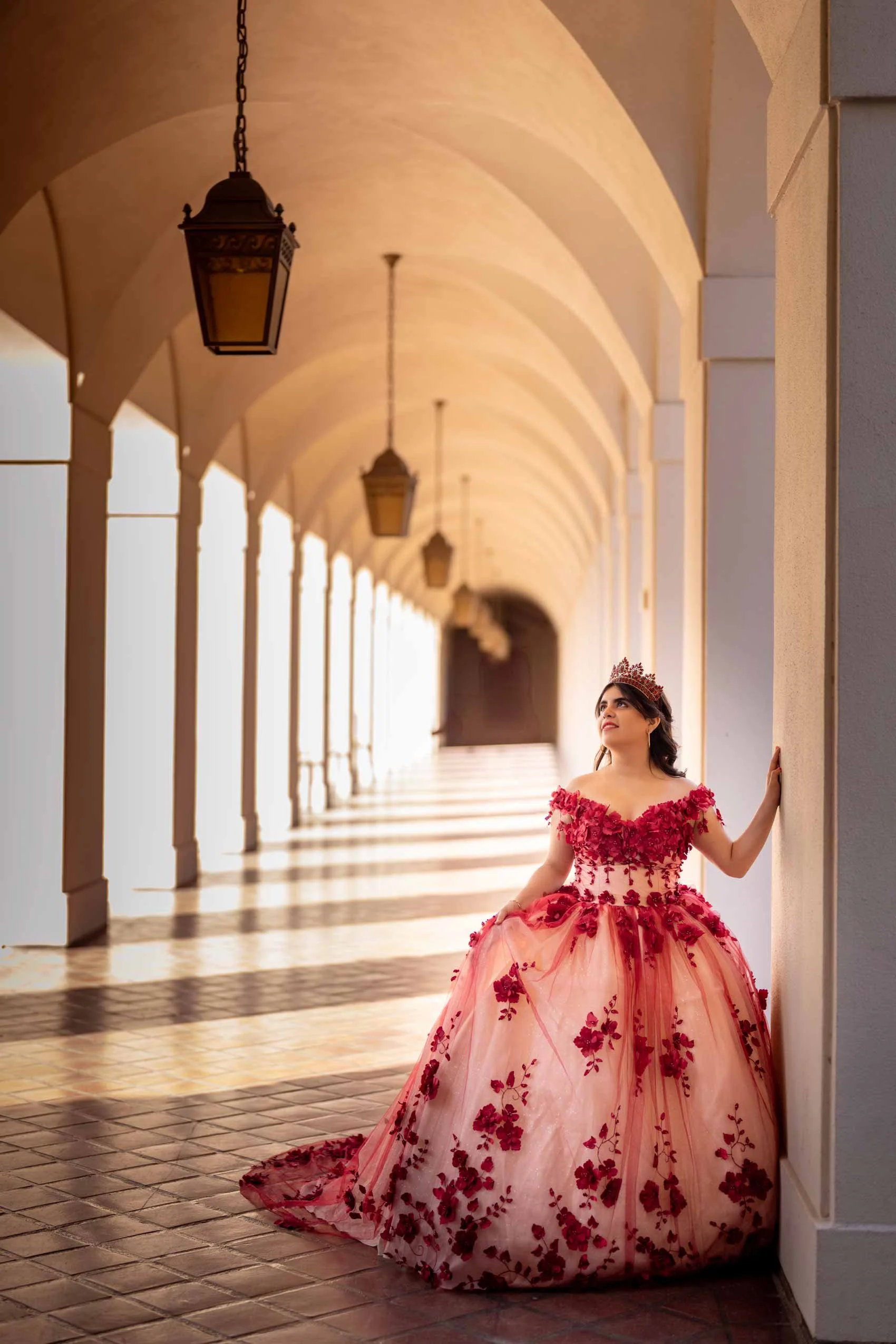 Young woman in a pink and red floral ball gown and tiara standing in an arched corridor with hanging lanterns.
