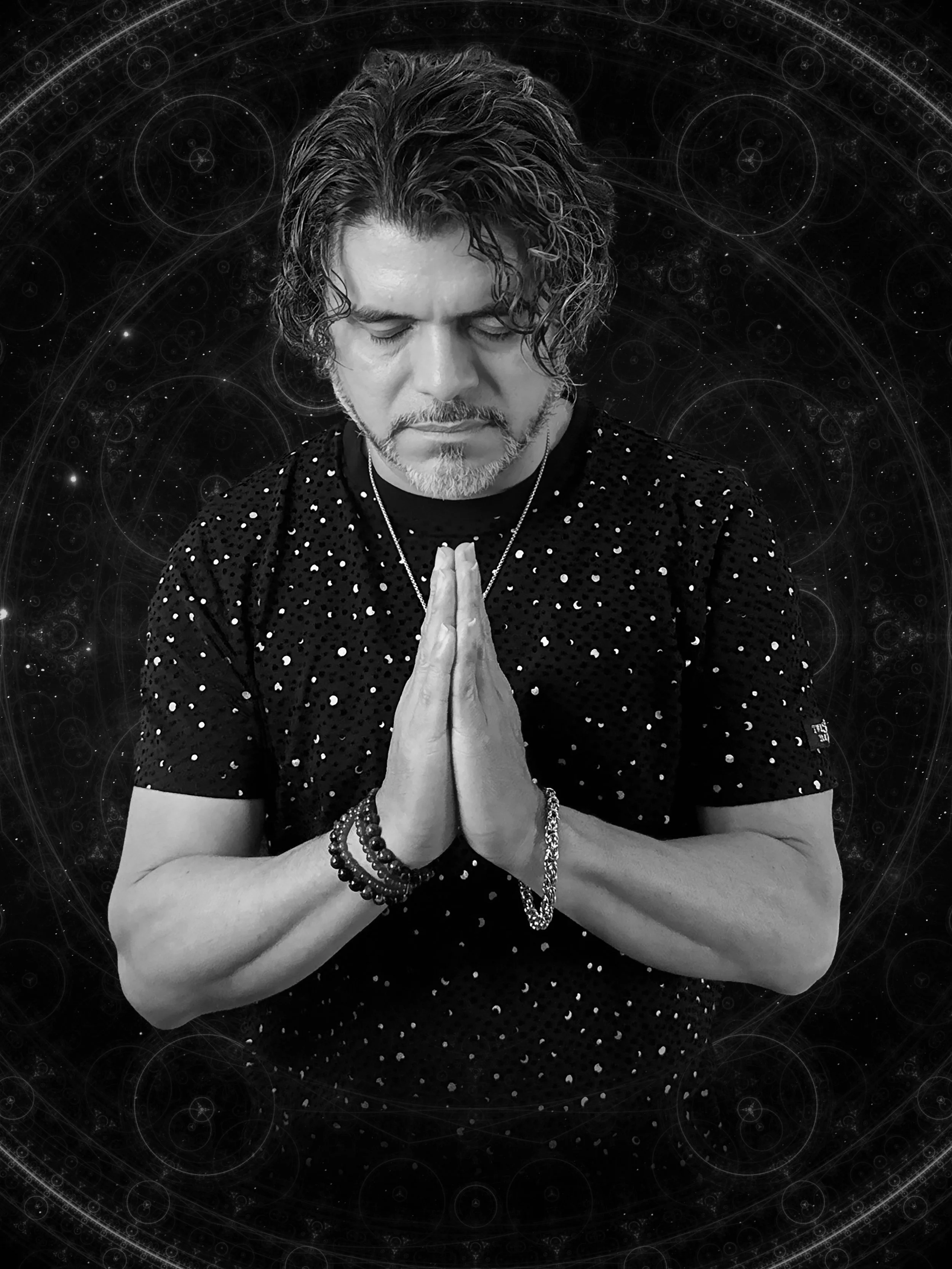 Black and white photo of a man with curly hair and a beard, with closed eyes and hands together in a prayer or meditation pose. The background features spiritual or cosmic patterns.