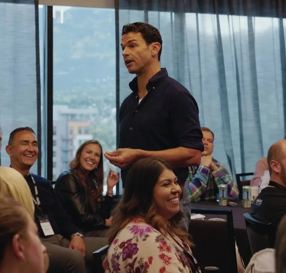 Man in dark shirt speaking at a conference, surrounded by smiling audience members in a room with large windows.