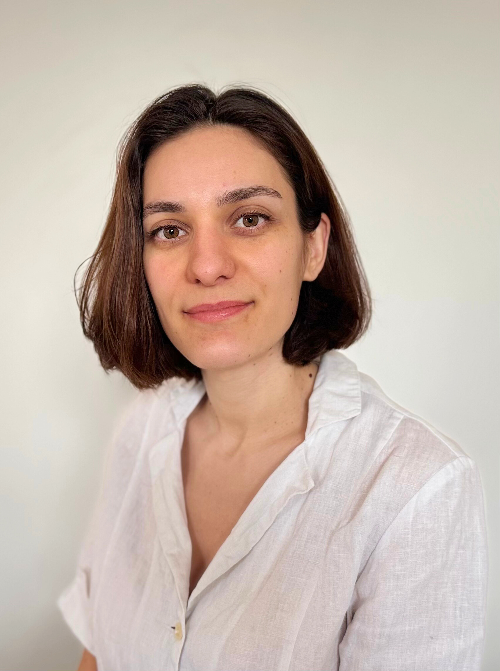 Jeune femme avec des cheveux bruns, portant une chemise blanche, regardant vers l'objectif avec un léger sourire, debout devant un mur blanc.