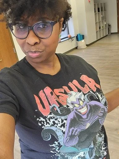A woman with short, curly hair and glasses taking a selfie indoors. She is wearing a black T-shirt with a graphic design of the villainous character Ursula from Disney's The Little Mermaid.