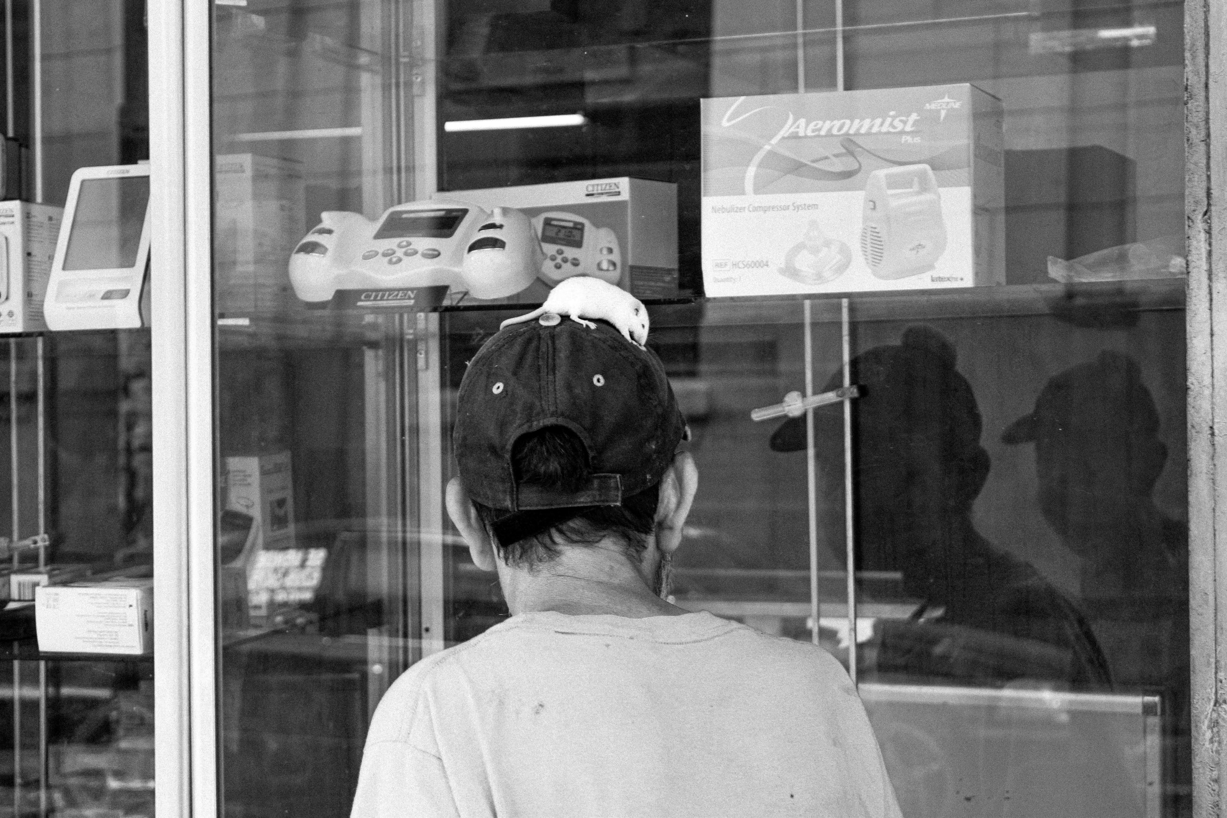 A man stands looking at the electrodomestic shop while walking with a rat on his head.