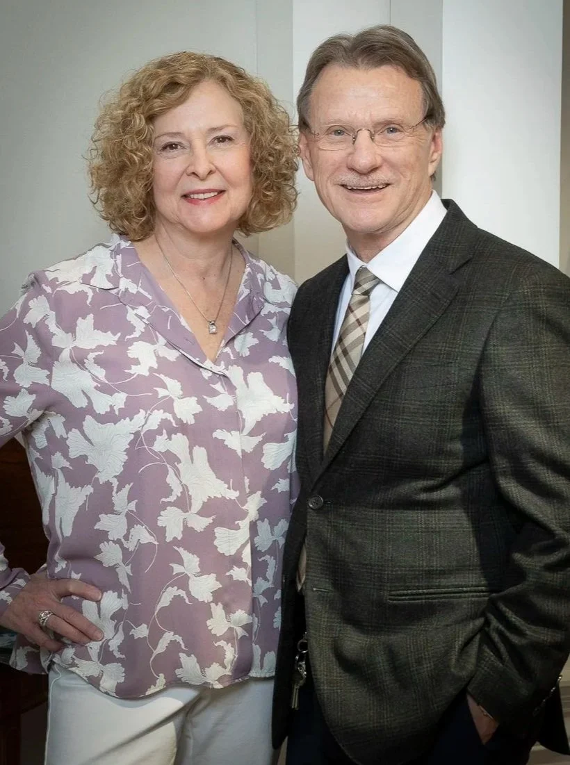 A smiling middle-aged woman with curly blonde hair wearing a purple floral blouse and a necklace, standing next to a smiling middle-aged man with glasses and short gray hair, dressed in a dark gray suit with a checkered tie.