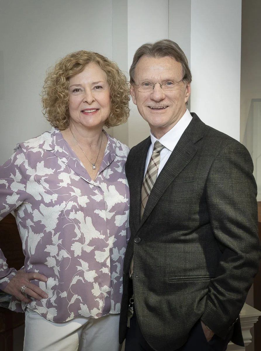 A smiling middle-aged woman with curly blonde hair wearing a lavender blouse with a white floral pattern, standing next to a smiling middle-aged man with glasses, wearing a dark blazer, white shirt, and patterned tie, both posing indoors in front of a white wall.