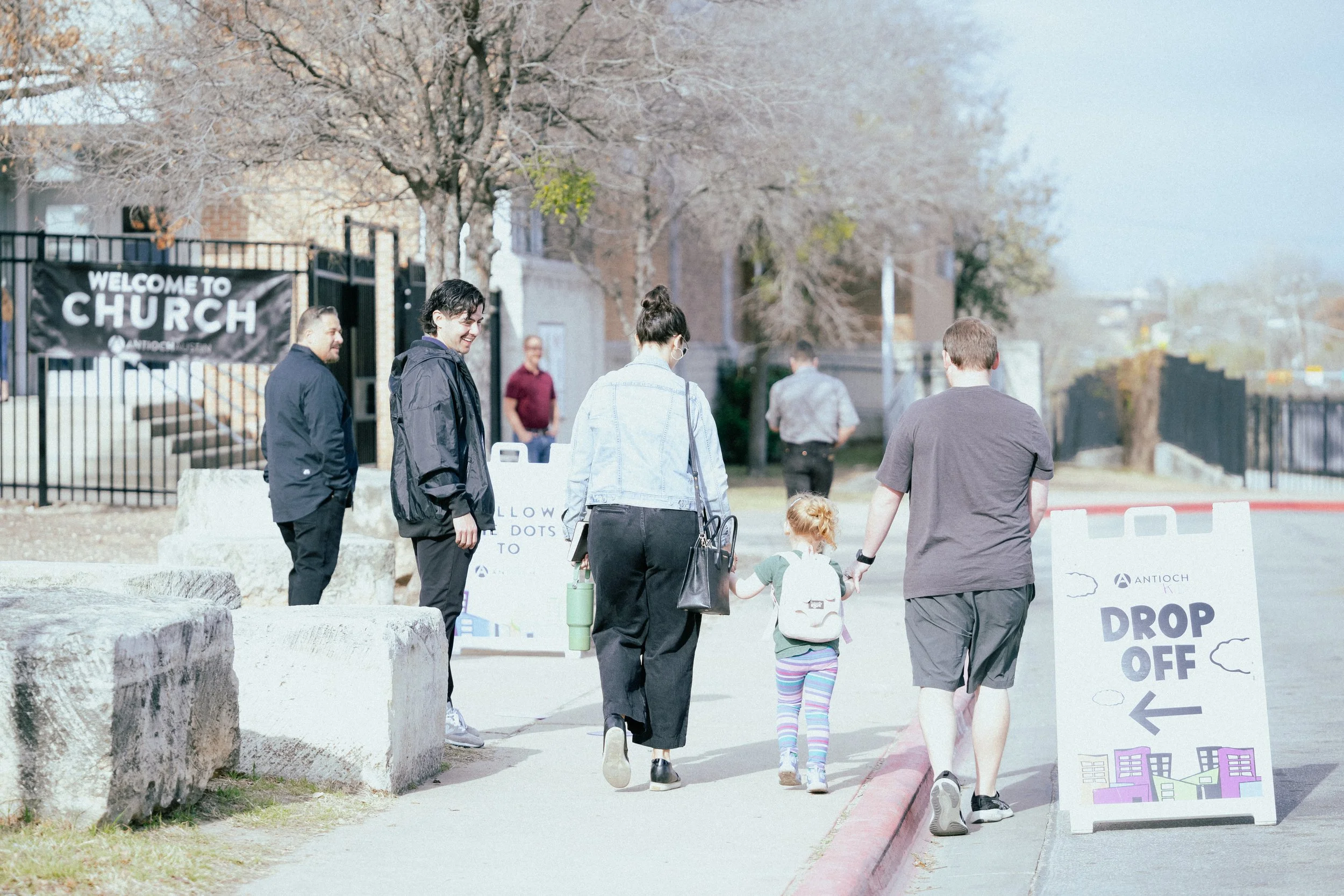 A group of people, including an adult with a backpack holding hands with children, walking toward a brick building with a sign offering free breakfast for students.