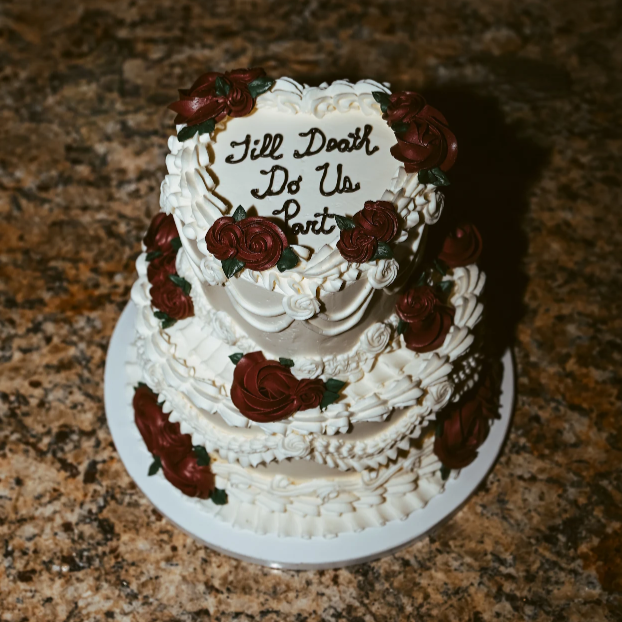 A tall, elegant cake decorated with blue and gray frosting flowers on a round, gold cake board, placed on a dark table.