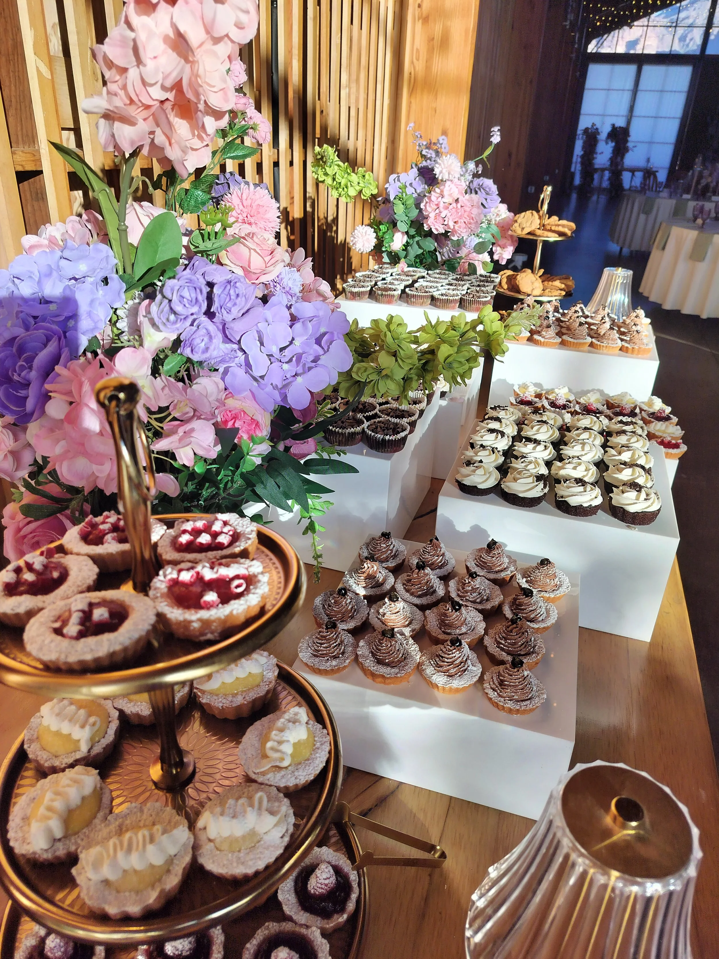 Assorted cupcakes and pastries arranged on white stands and trays, with decorative pink, purple, and green floral arrangements, in a setting with wooden paneling and soft lighting.