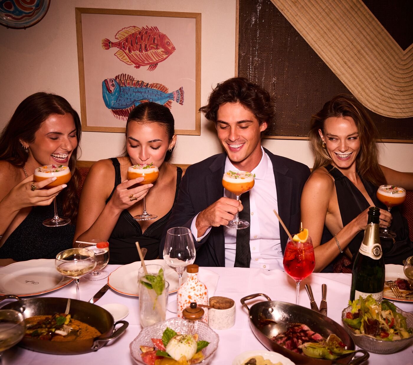 Four friends smiling over colorful cocktails and a dinner spread at a restaurant