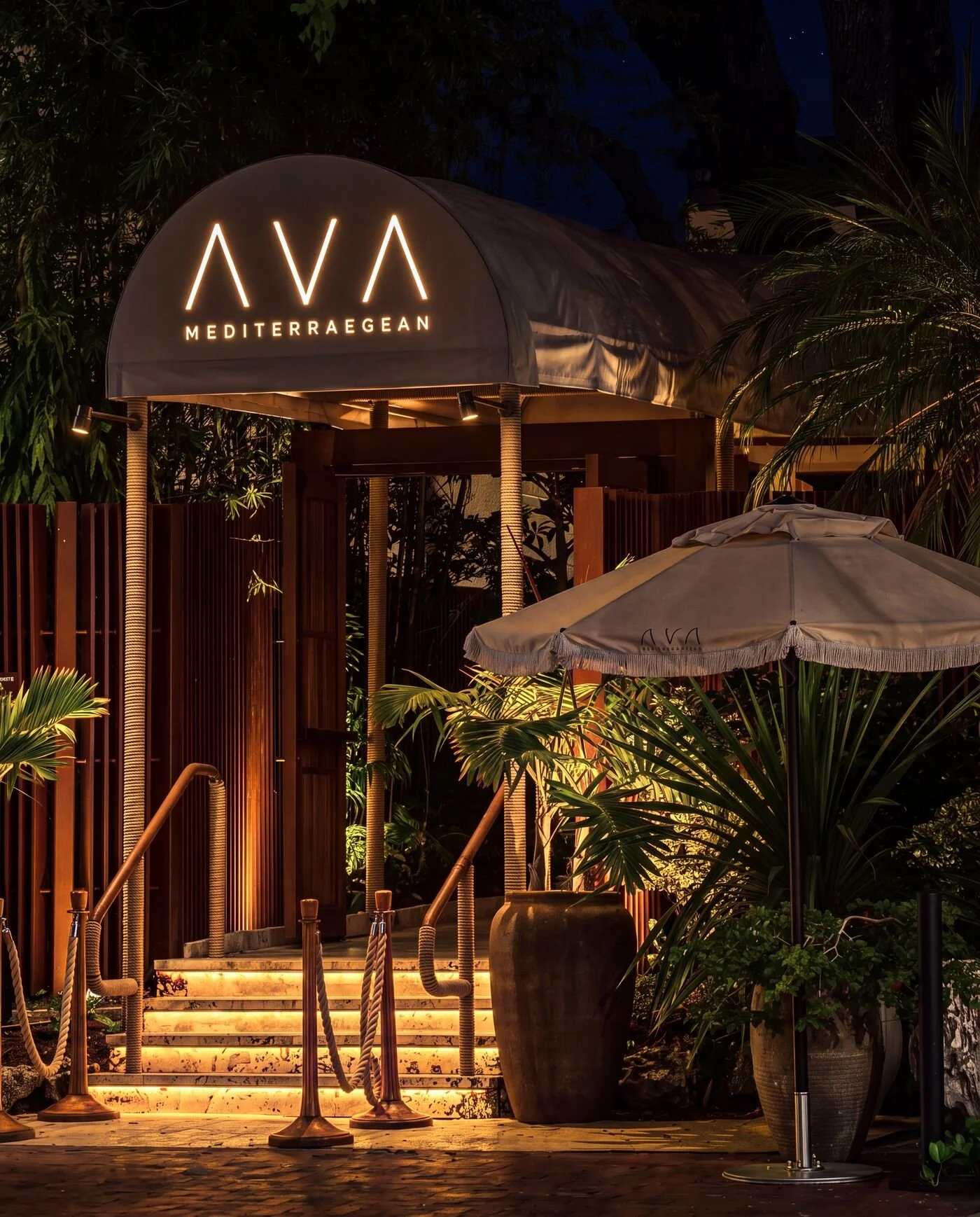 Nighttime entrance to AVA Mediterranean restaurant surrounded by tropical foliage, with illuminated stairs, canopy, and patio umbrella.