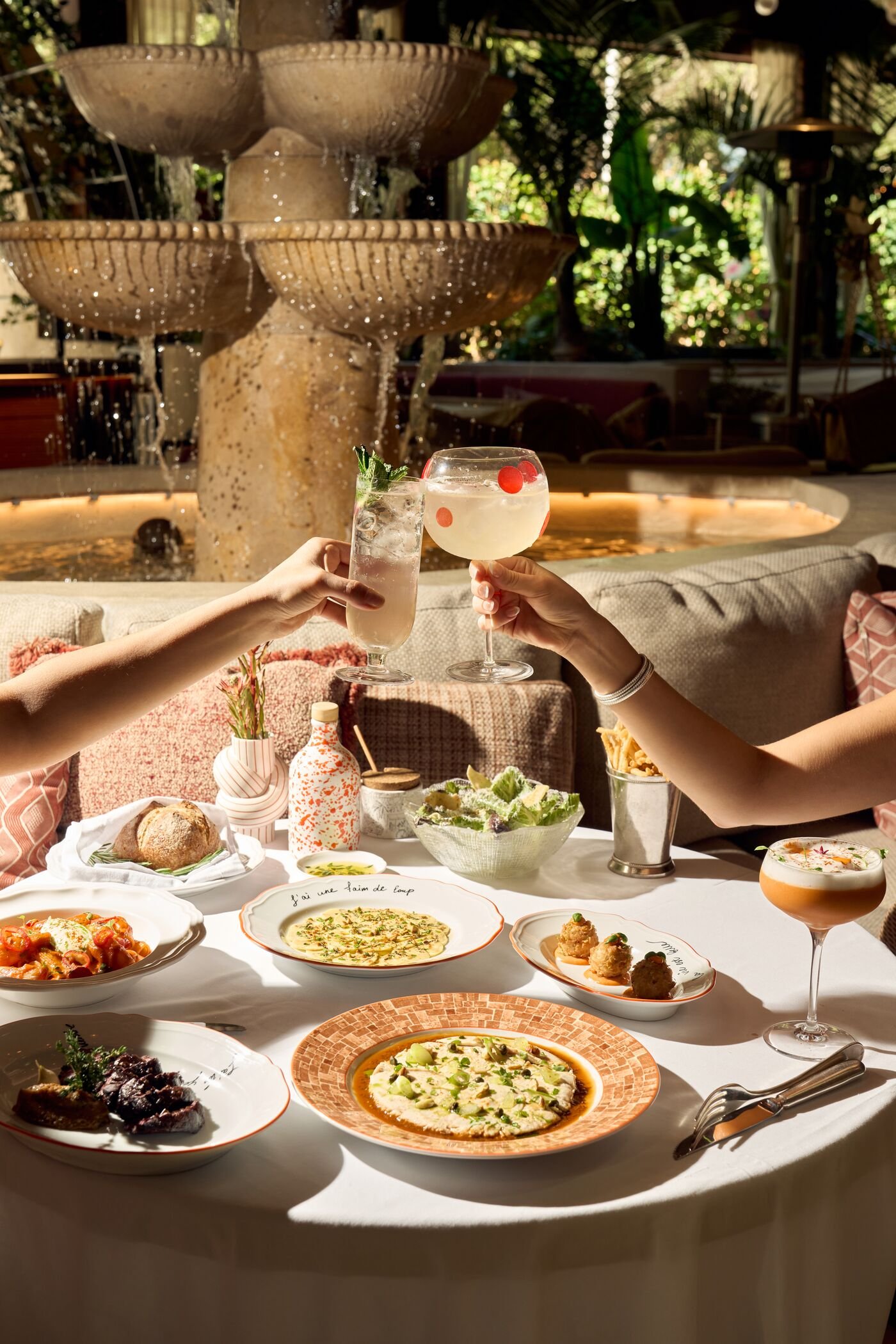 A table set with various dishes including pizza, pasta, and salads, with two hands holding glasses of cocktails, in front of a fountain and lush greenery.