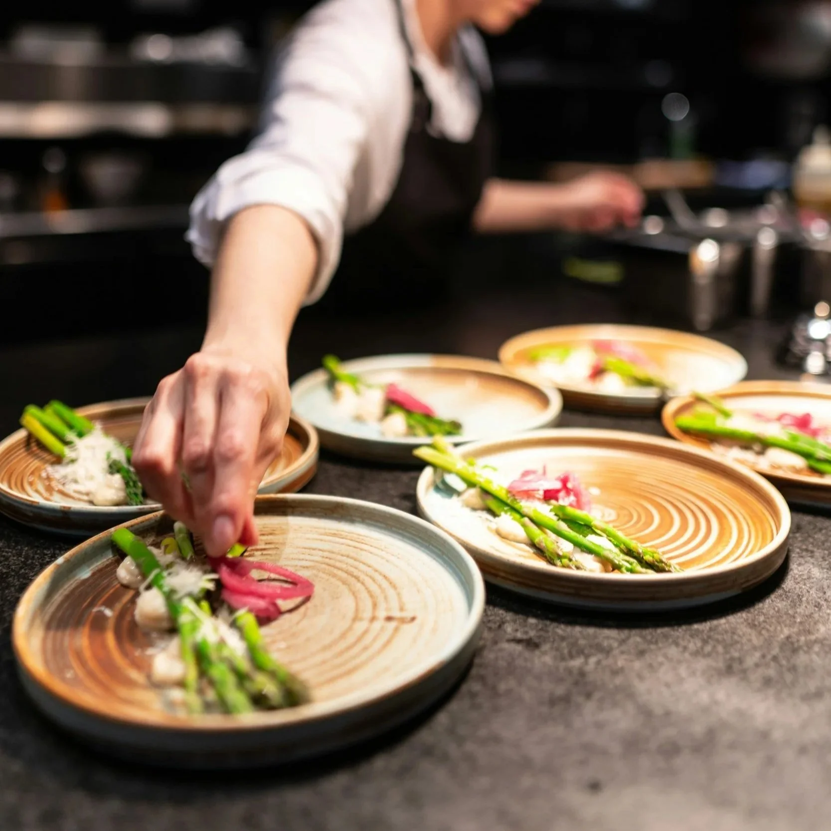 A chef garnishing small plates of food with asparagus, pink pickled onions, and other ingredients in a restaurant kitchen.