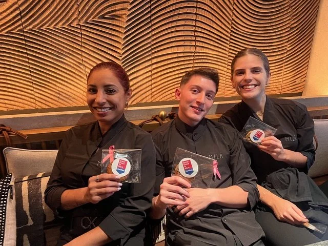 Three women in black shirts holding small round objects with ribbon symbols, sitting on a bench and smiling, in front of a wooden wall with carved patterns.