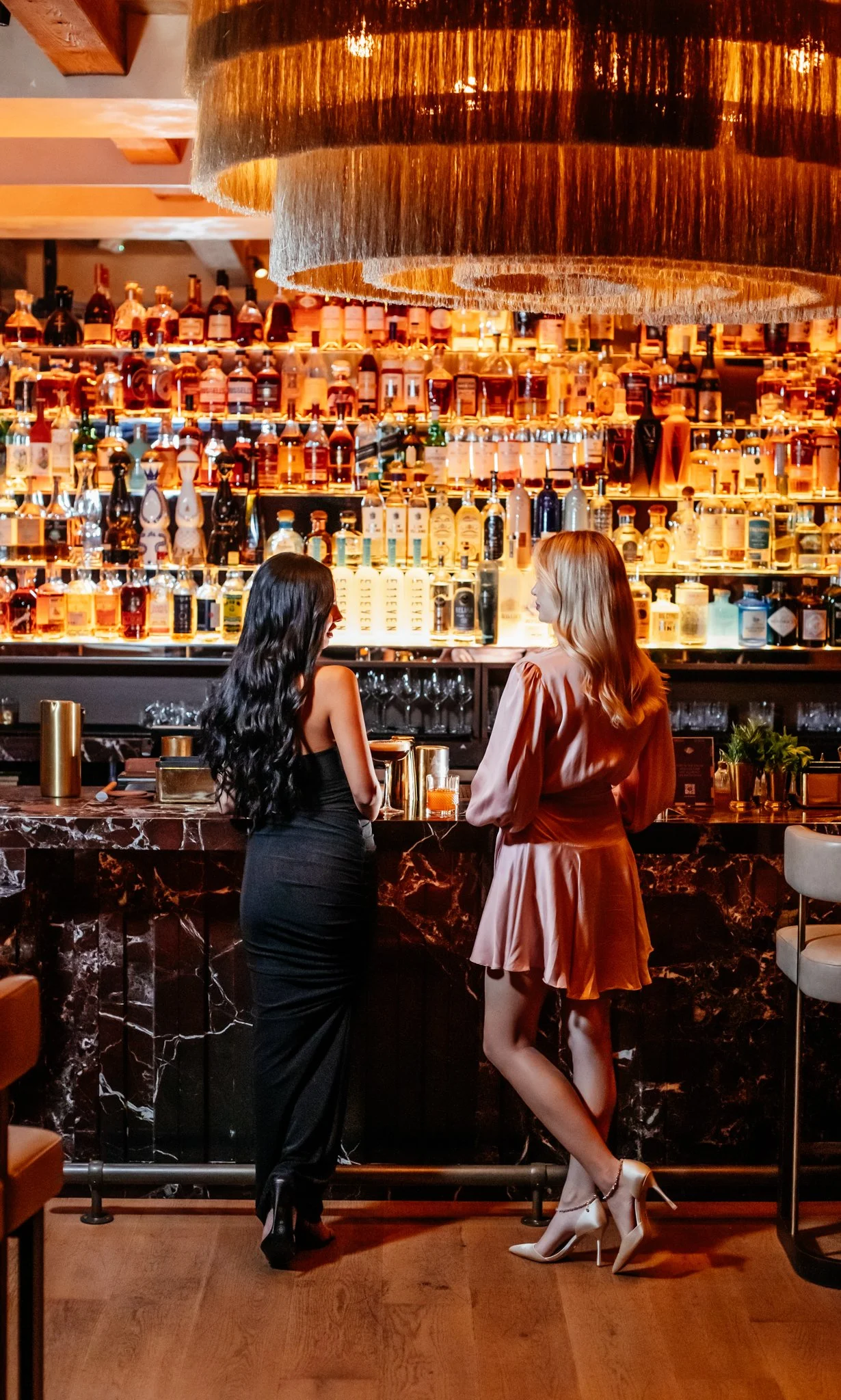 Two elegant women at the exclusive members only section, where they are waiting for their cocktails to be made at the elegant bar.