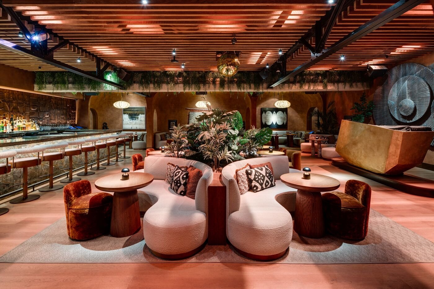 Interior of a stylish lounge bar with a curved sofa, patterned cushions, surrounded by small tables and chairs, plants, warm lighting, and a bar area with bottles on the left.