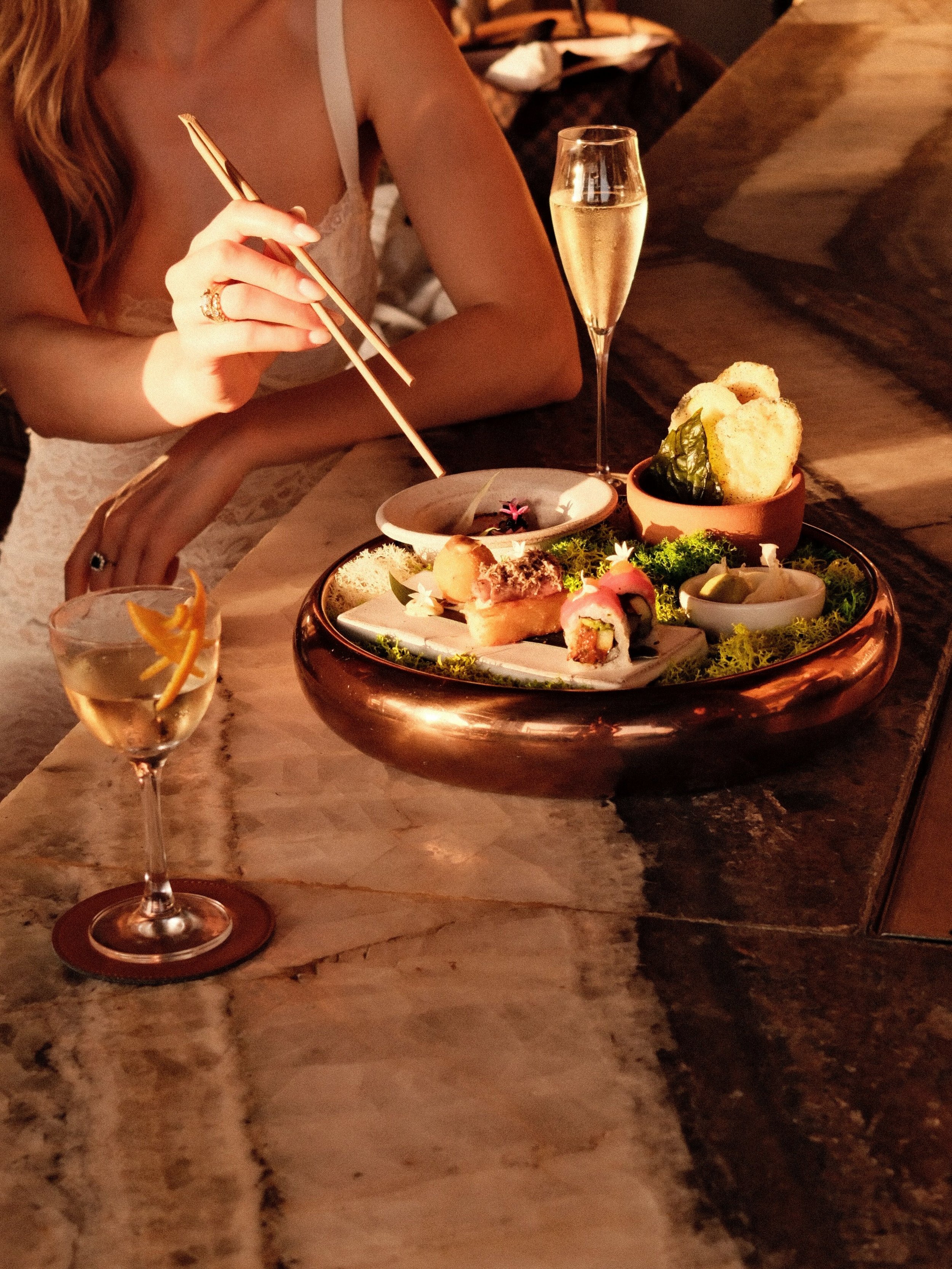 A woman is sitting at a table with a variety of sushi and Japanese food, a glass of champagne, and a cocktail with an orange garnish. The setting appears to be in a dimly lit restaurant.
