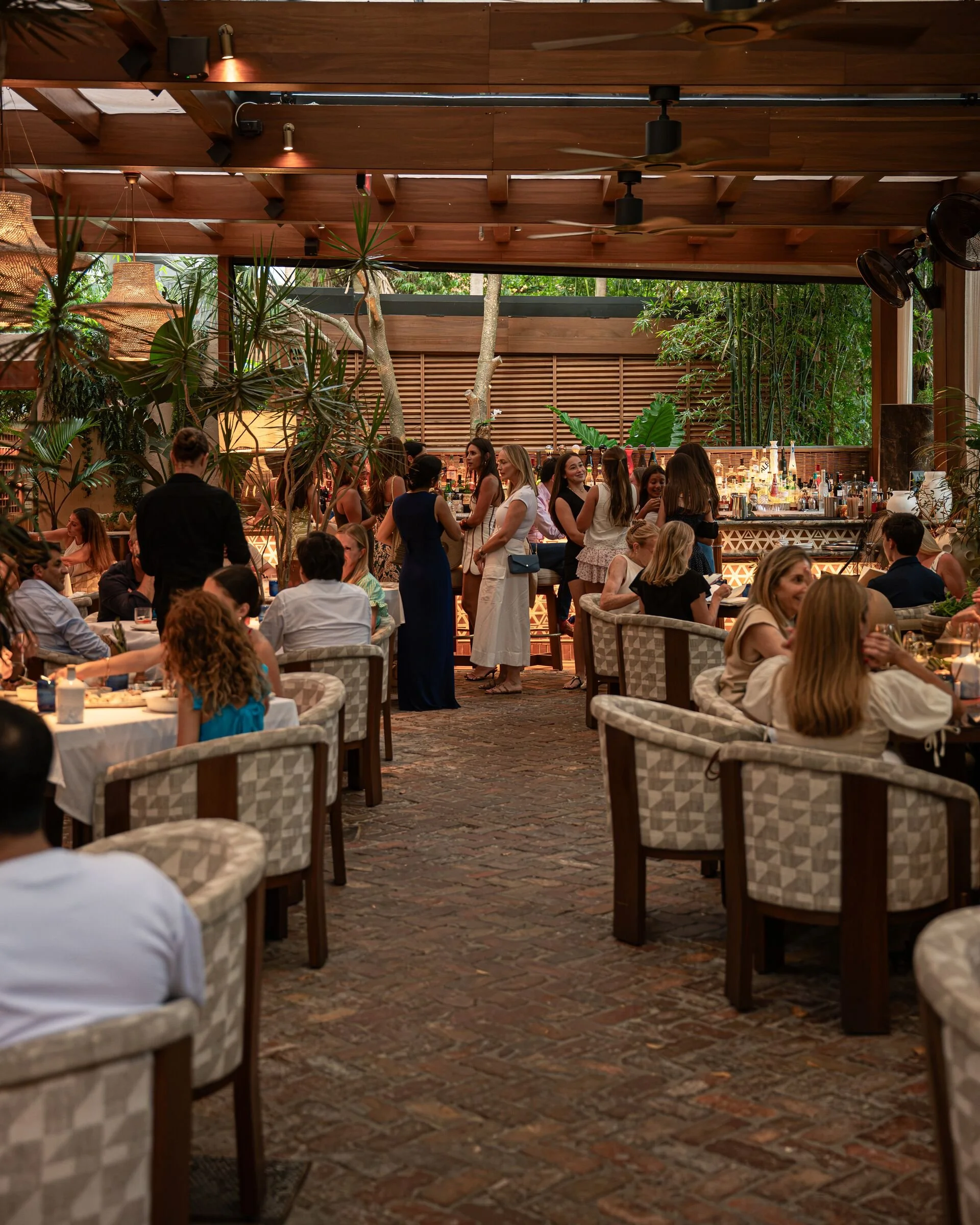 Busy tropical restaurant with diners, a well-stocked bar, lush plants, woven pendant lights, and exposed wooden beams.