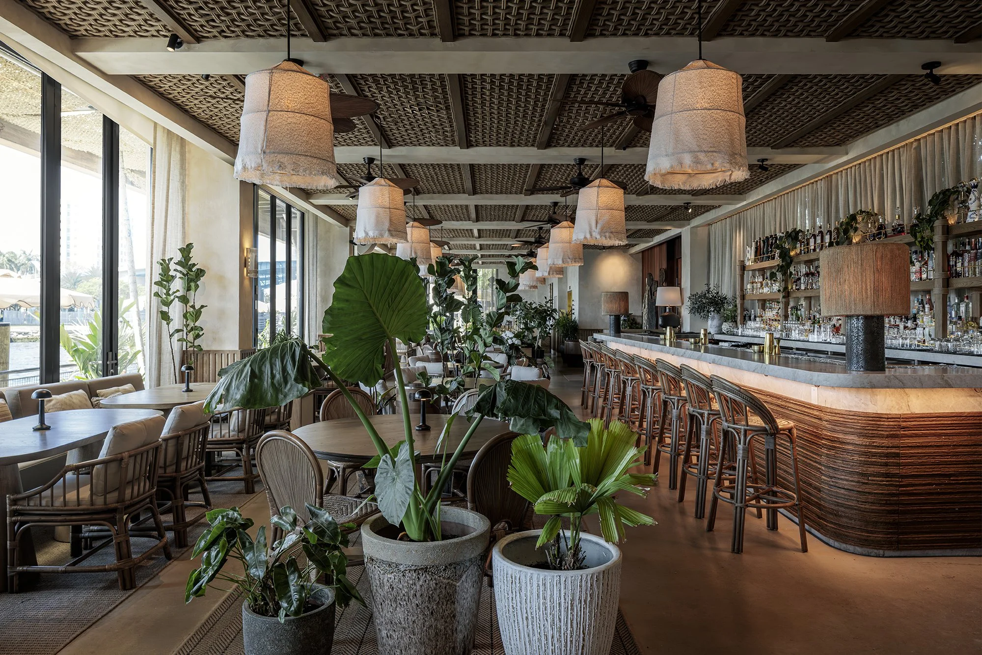 The restaurant dining room of the inside of CASA NEOS, with tables, greenery, and a bar on the right side.