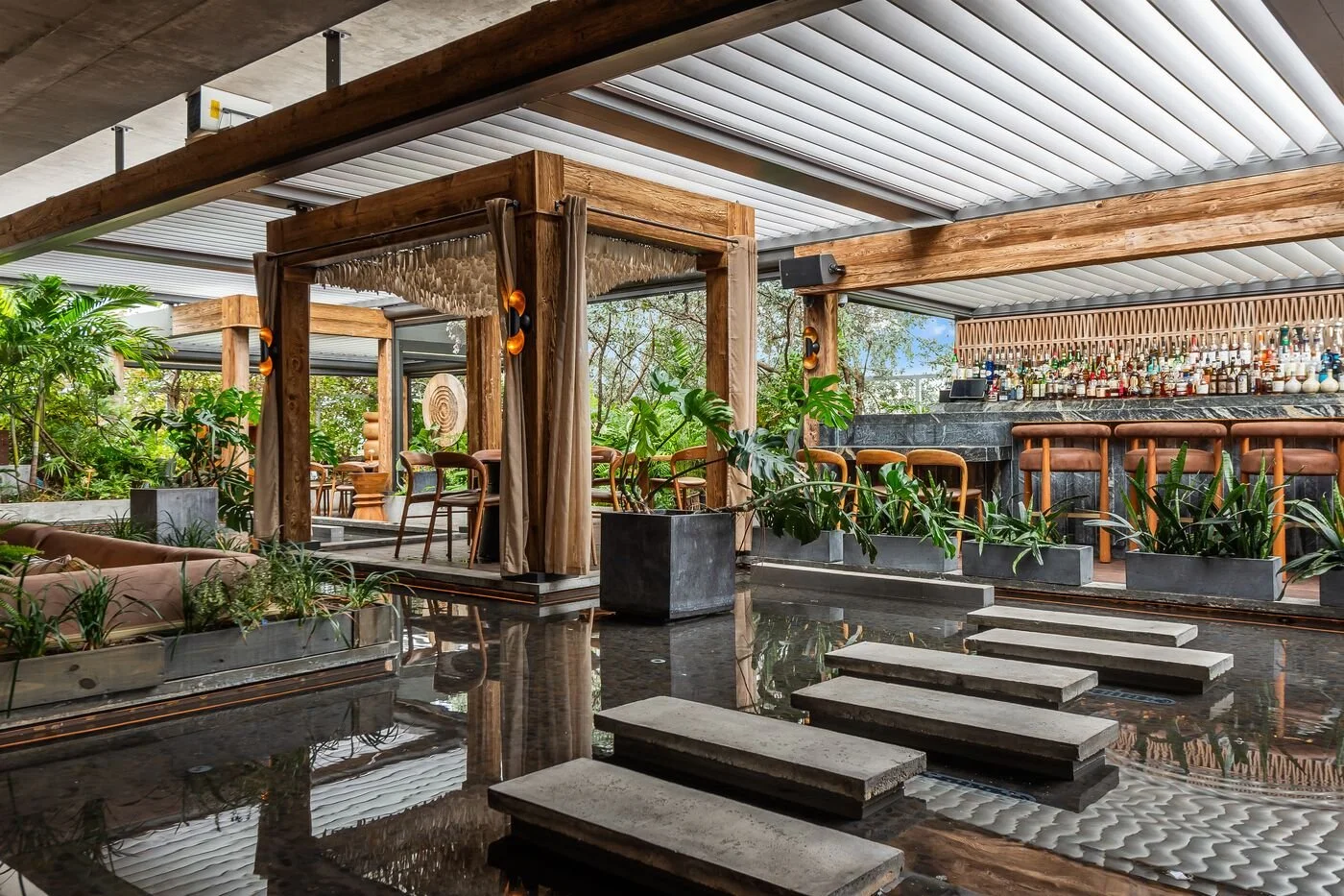 A stylish indoor lounge area with a bar, wooden furniture, and greenery, featuring a modern design with plants, steps, and a bar counter in the background.