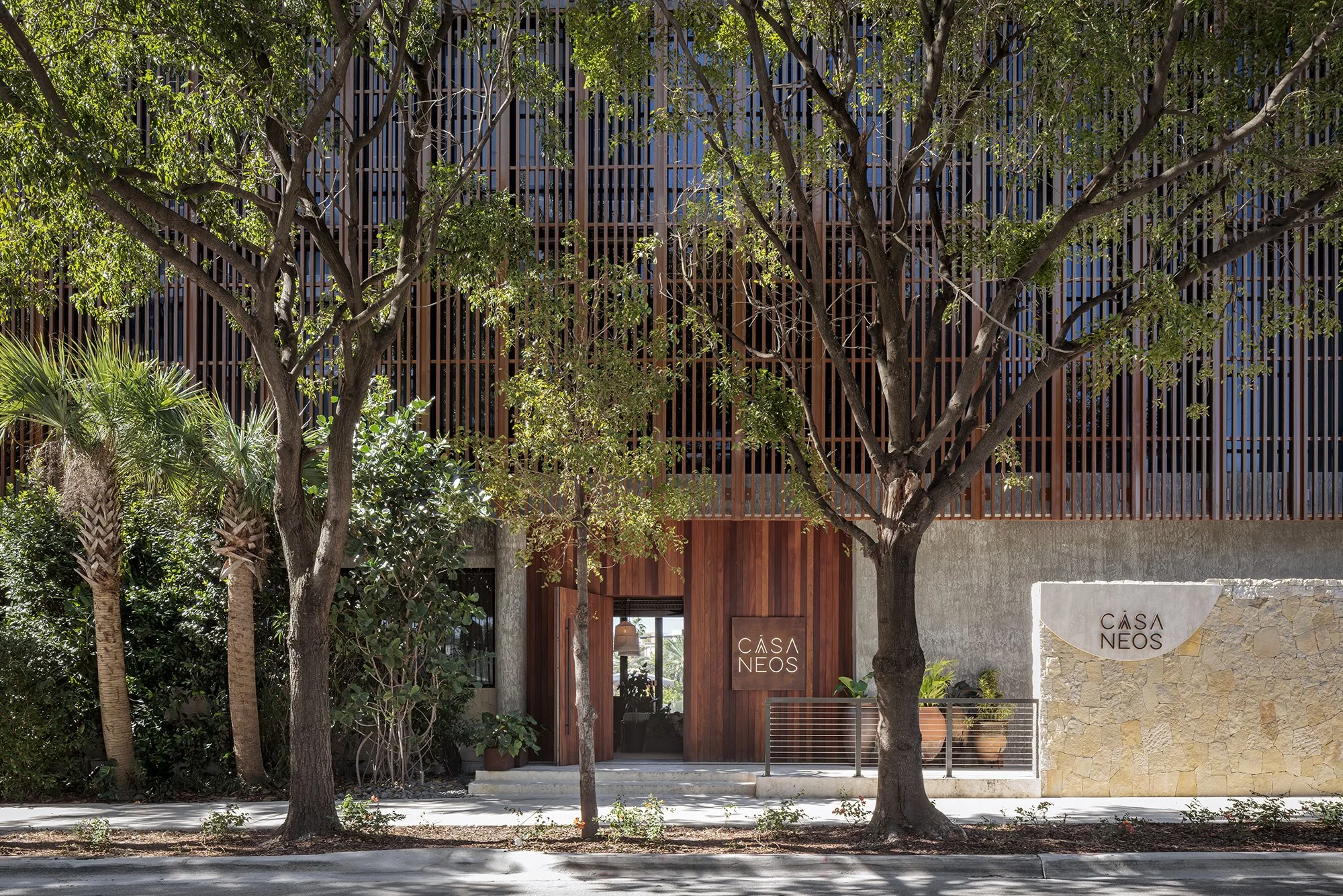 Entrance of CASA NEOS, with a beach exterior decoration, including a wood-styled door and a stone slab with CASA NEOS logo on the top.