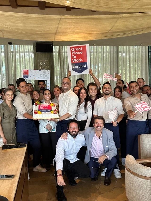 The riviera dining group team celebrating being certified as a "Great Place to Work", having a cake to celebrate and festivity decorations.
