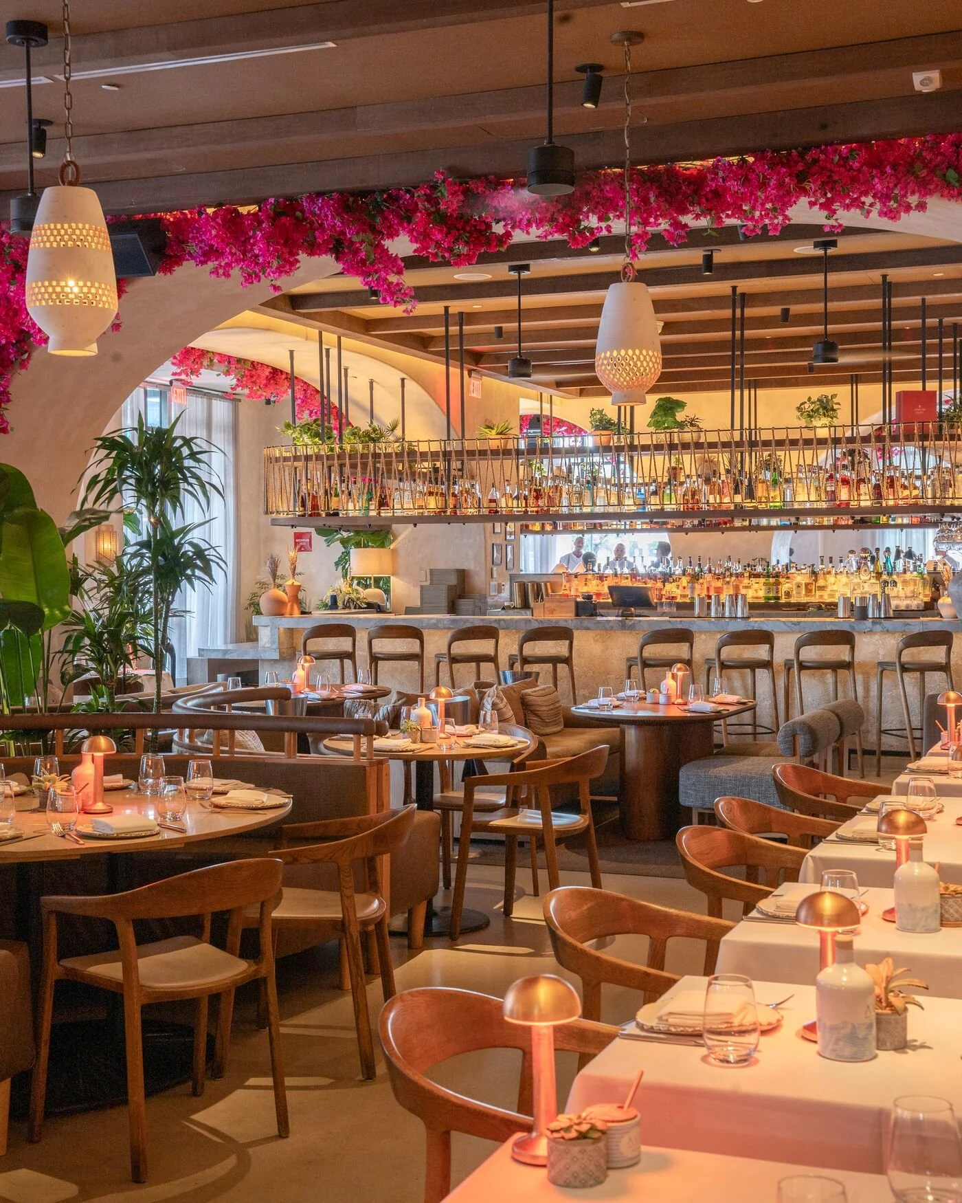 Elegant restaurant interior with wooden accents, pink flower decorations, various plants, and neatly arranged tables with lit candles, glasses, and plates.
