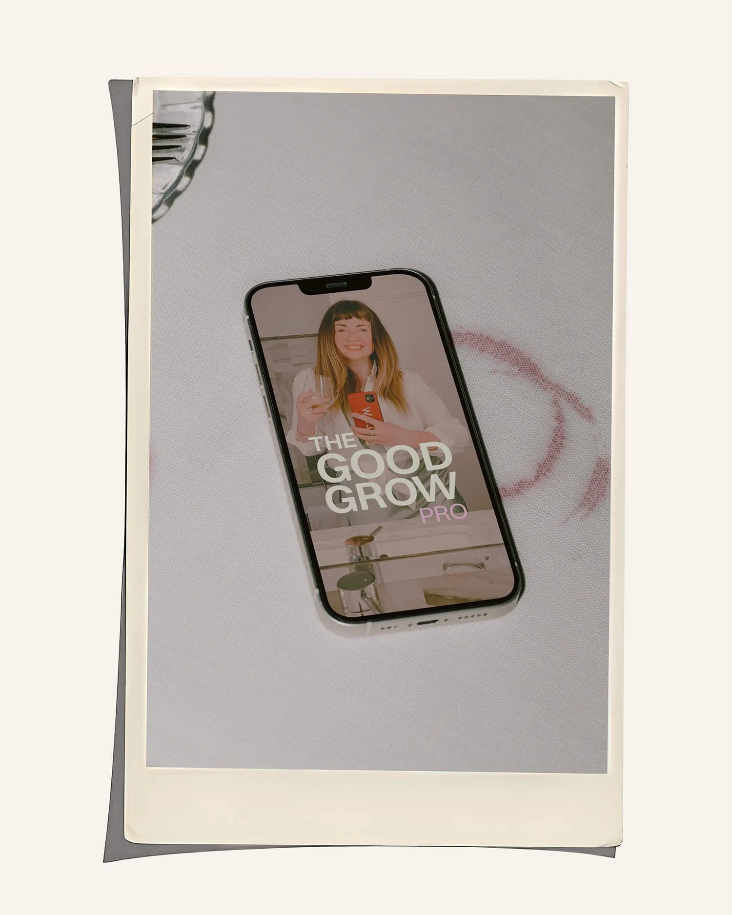 Smartphone on a white surface displaying a woman taking a selfie with the text "The Good Grow PRO" on the screen.
