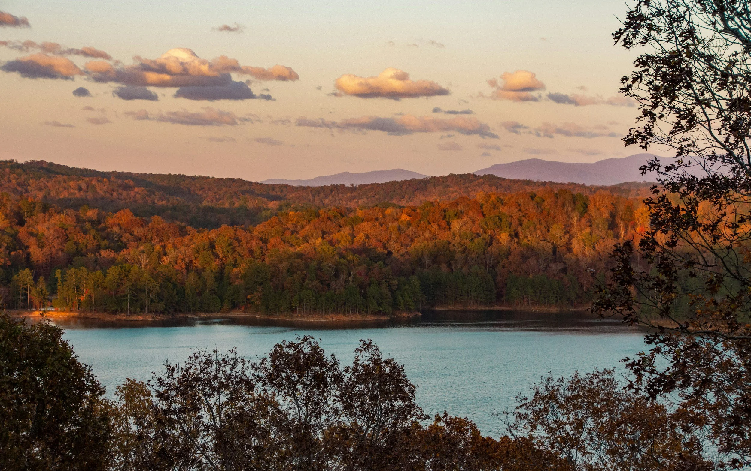 Scenic view of a lake surrounded by trees with autumn foliage, with hills and mountains in the background under a sky with scattered clouds. Located in North Georgia offering cleaning services in Gilmer, Pickens, Fannin, counties for airbnbs