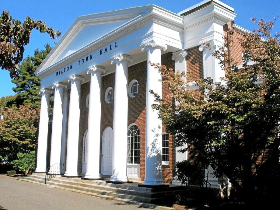 Front view of a classical-style building with large white columns and brick facade, labeled "Wilton Town Hall." The building is surrounded by trees.