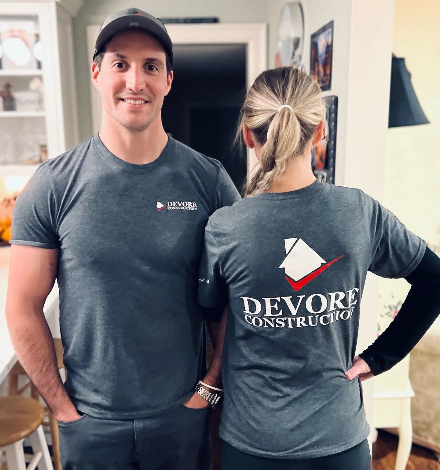 Two people wearing 'Devore Construction' branded gray t-shirts, one facing front and one showing the back, in a home setting.