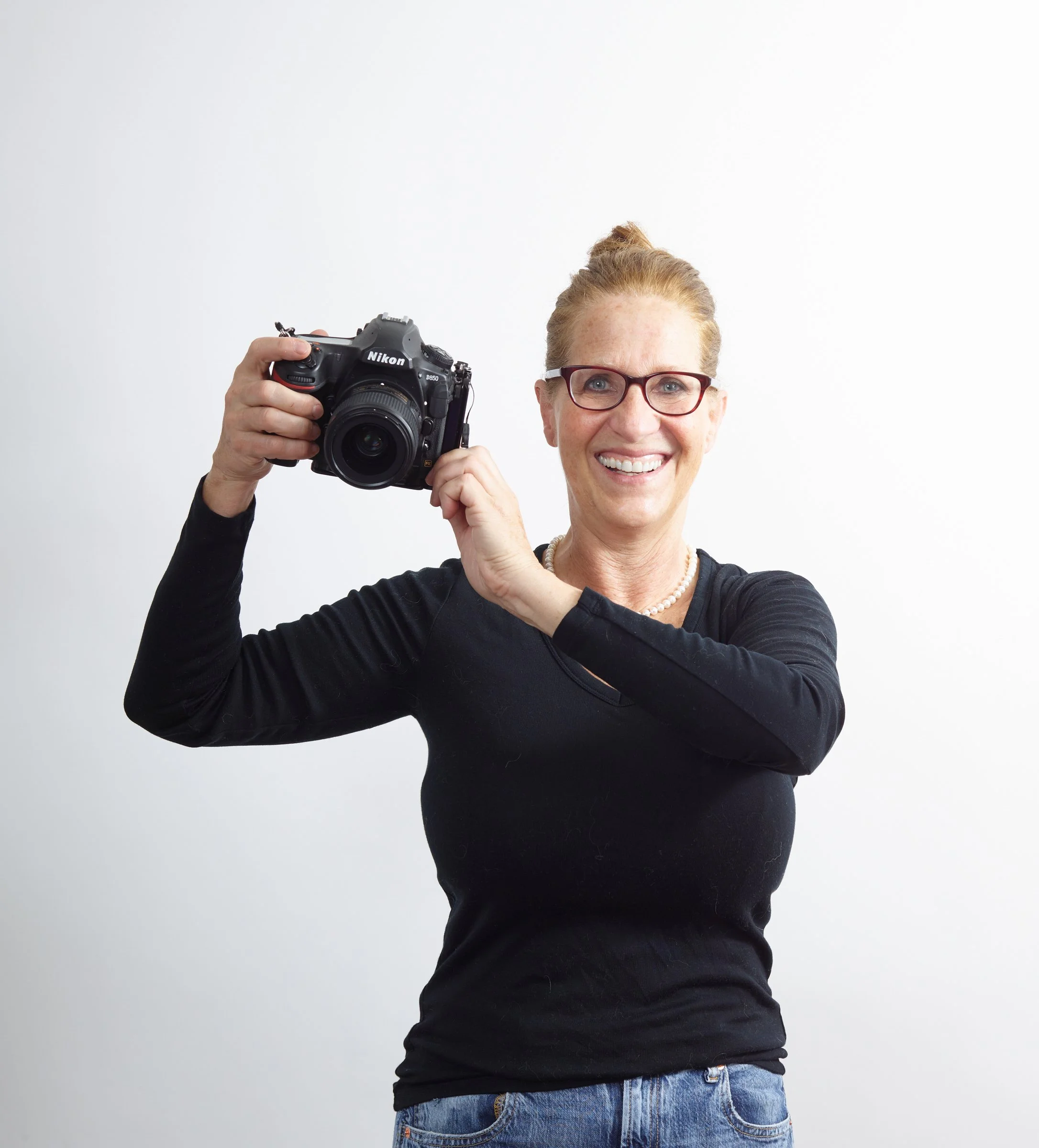 Judy Doherty, food photographer, self-portrait, with glasses holding a Nikon camera, standing against a white background.