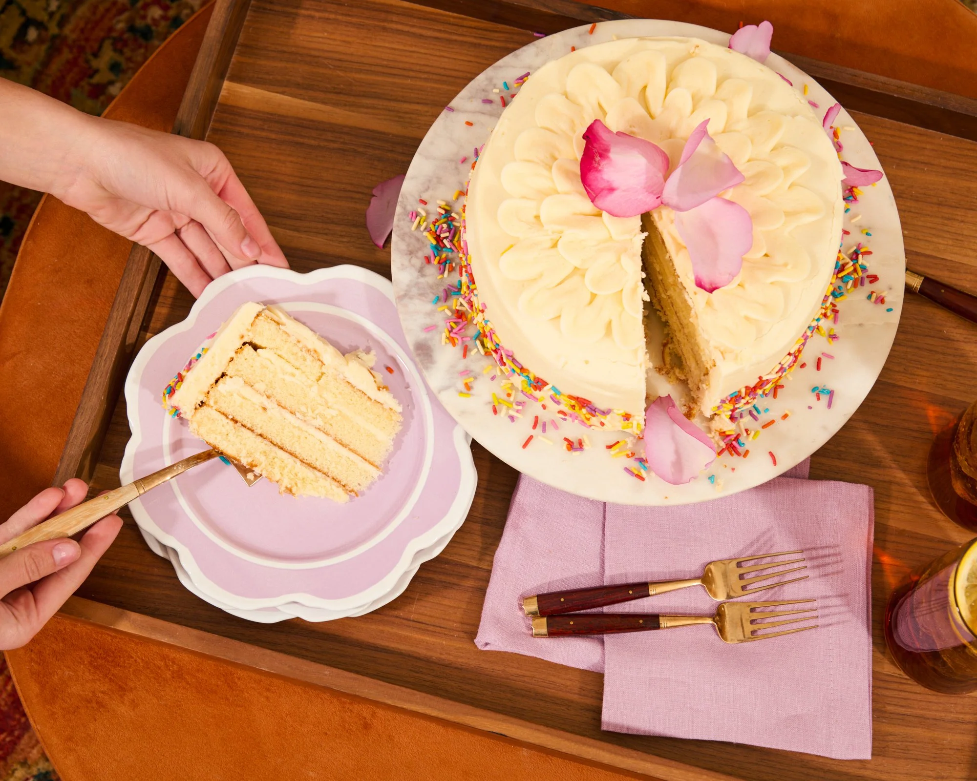 A round birthday cake with white frosting, colorful sprinkles, and pink flower petals on top. A slice is being cut from the cake, revealing yellow cake layers. The cake is on a white marble cake stand. There are two small forks and pink napkin undern
